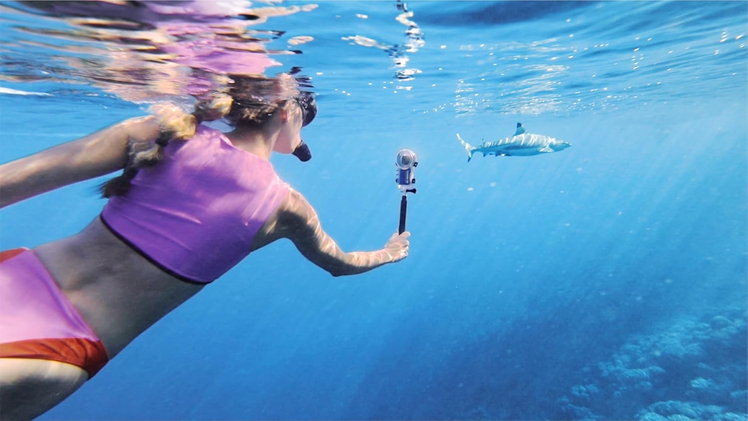 Mujer haciendo esnórquel, con cámara en un palo selfie, frente a un tiburón en agua azul clara.