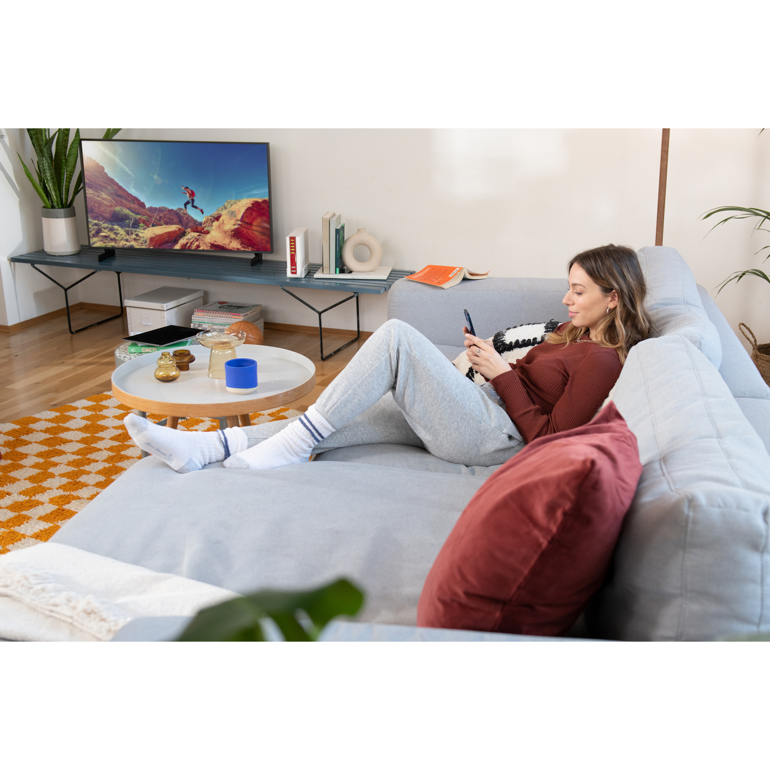 Femme sur canapé, téléphone, TV. Tapis jaune, table bleue, plante en arrière-plan.