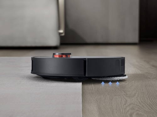 Czarny robot sprzątający czyszczący podłogę. Podłoga ma sekcje drewniane i dywanowe.