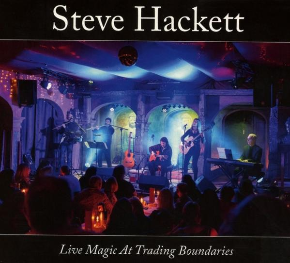 Albumcover: Steve Hackett Live Magic At Trading Boundaries. Band auf der Bühne mit Lichtern. Publikum anwesend.