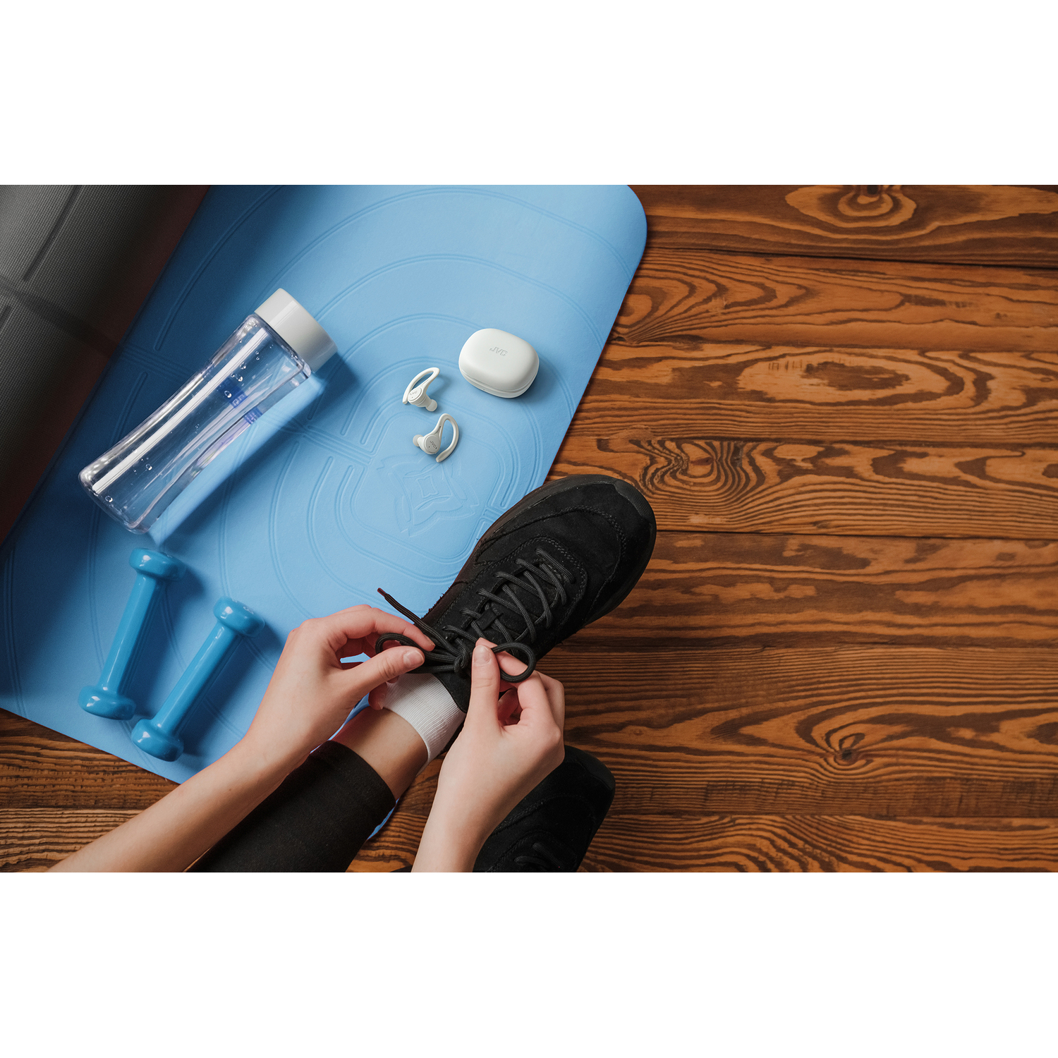 Une personne lace une chaussure noire sur un sol en bois avec un tapis bleu, des haltères et une gourde.
