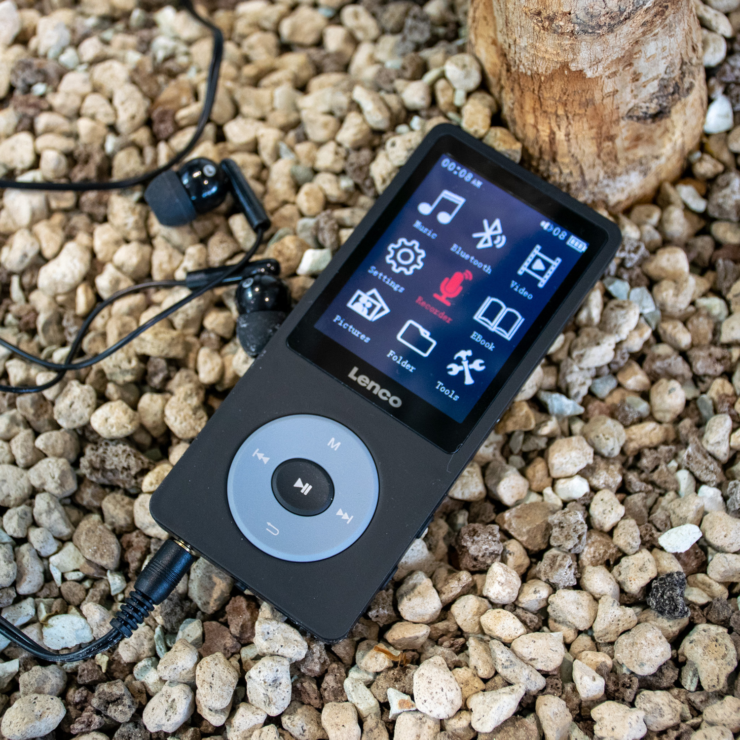 A black MP3 player with a screen, and headphones. It is on a bed of small, brown pebbles.