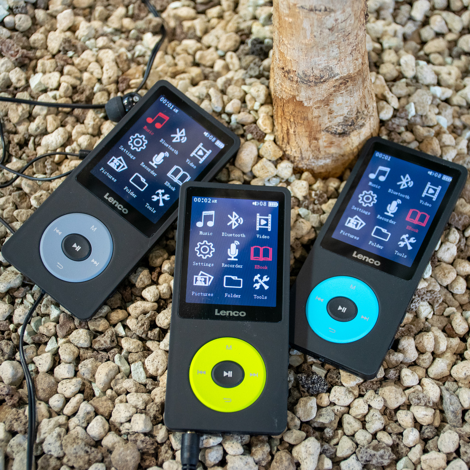 Three black Lenco MP3 players on a surface of small stones. Each has a screen and a different color button.