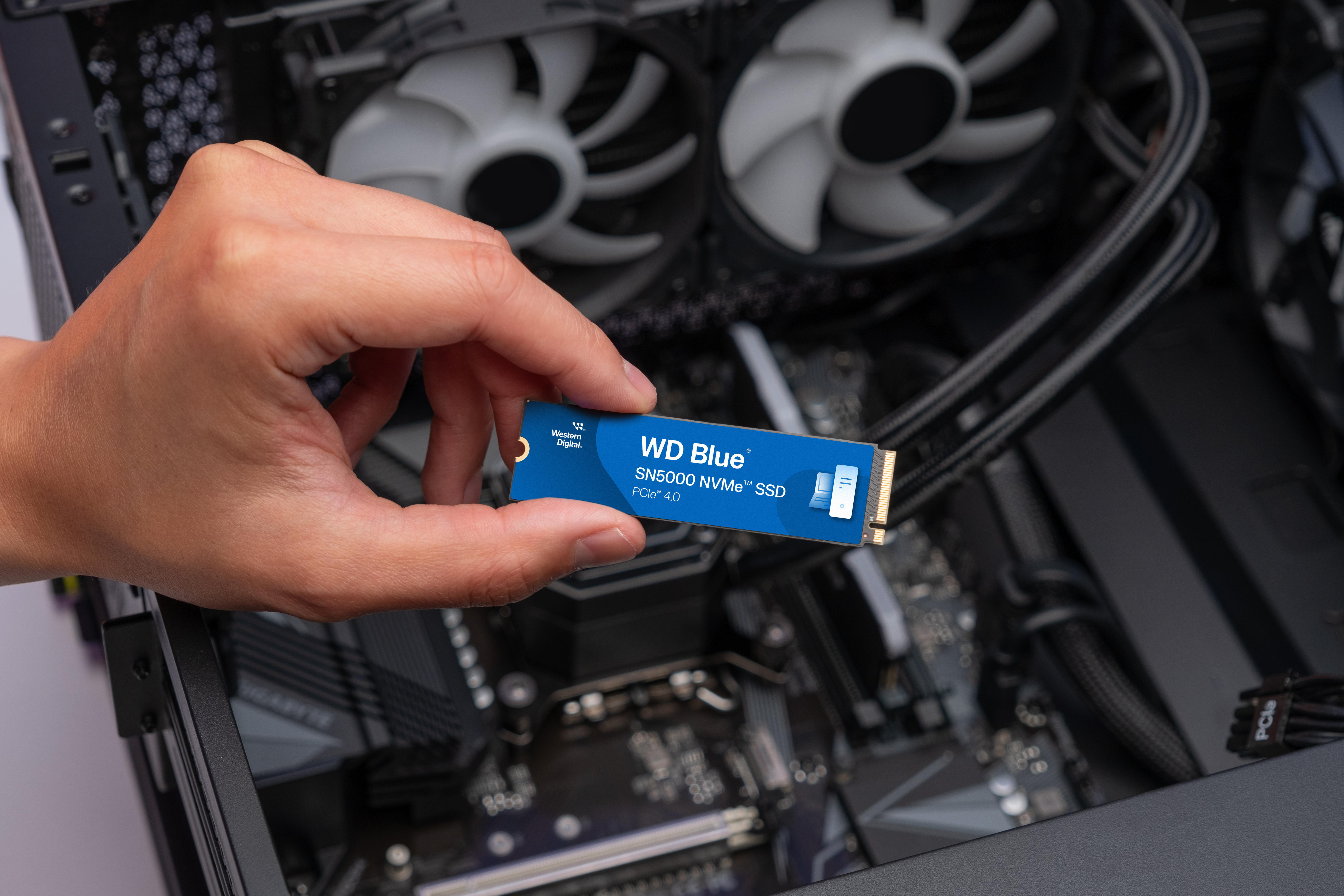 Eine Hand, die eine blaue WD_Blue SN5000 NVMe SSD über einem Computer hält.