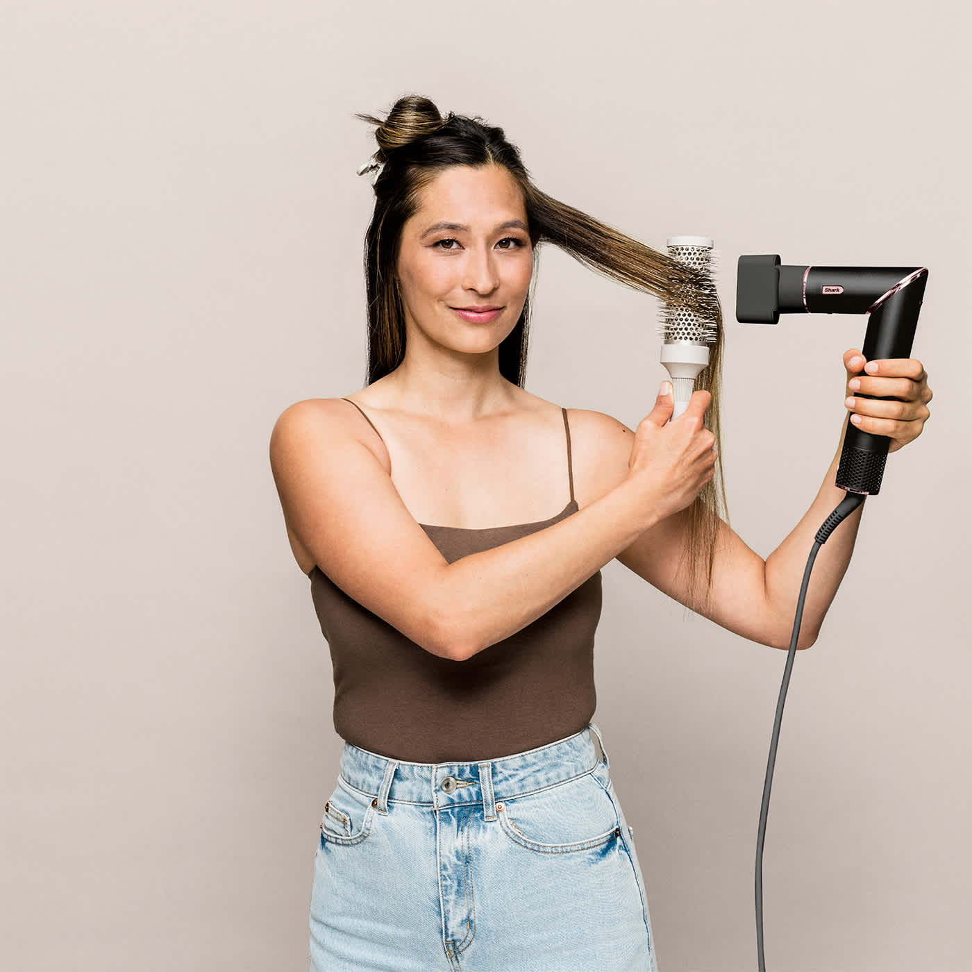 Eine Frau stylt ihre Haare mit einem Fön und einer Rundbürste. Sie trägt ein braunes Oberteil und Jeans.