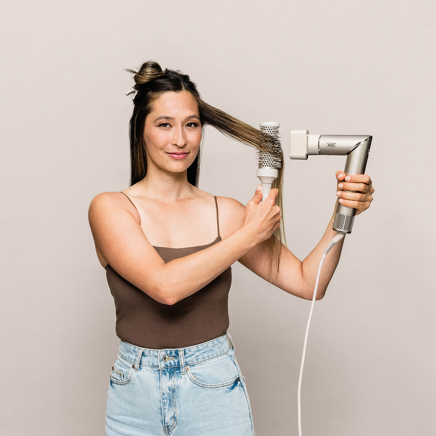 Frau, die ihr Haar mit einem silbernen und weißen Haartrockner stylt. Sie trägt ein braunes Tanktop und Jeans.
