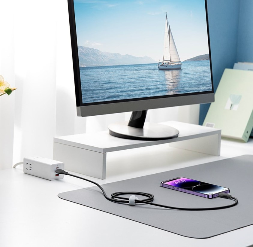Beyaz bir stand üzerinde bilgisayar monitörü, şarj cihazına bağlı. Masada telefon.