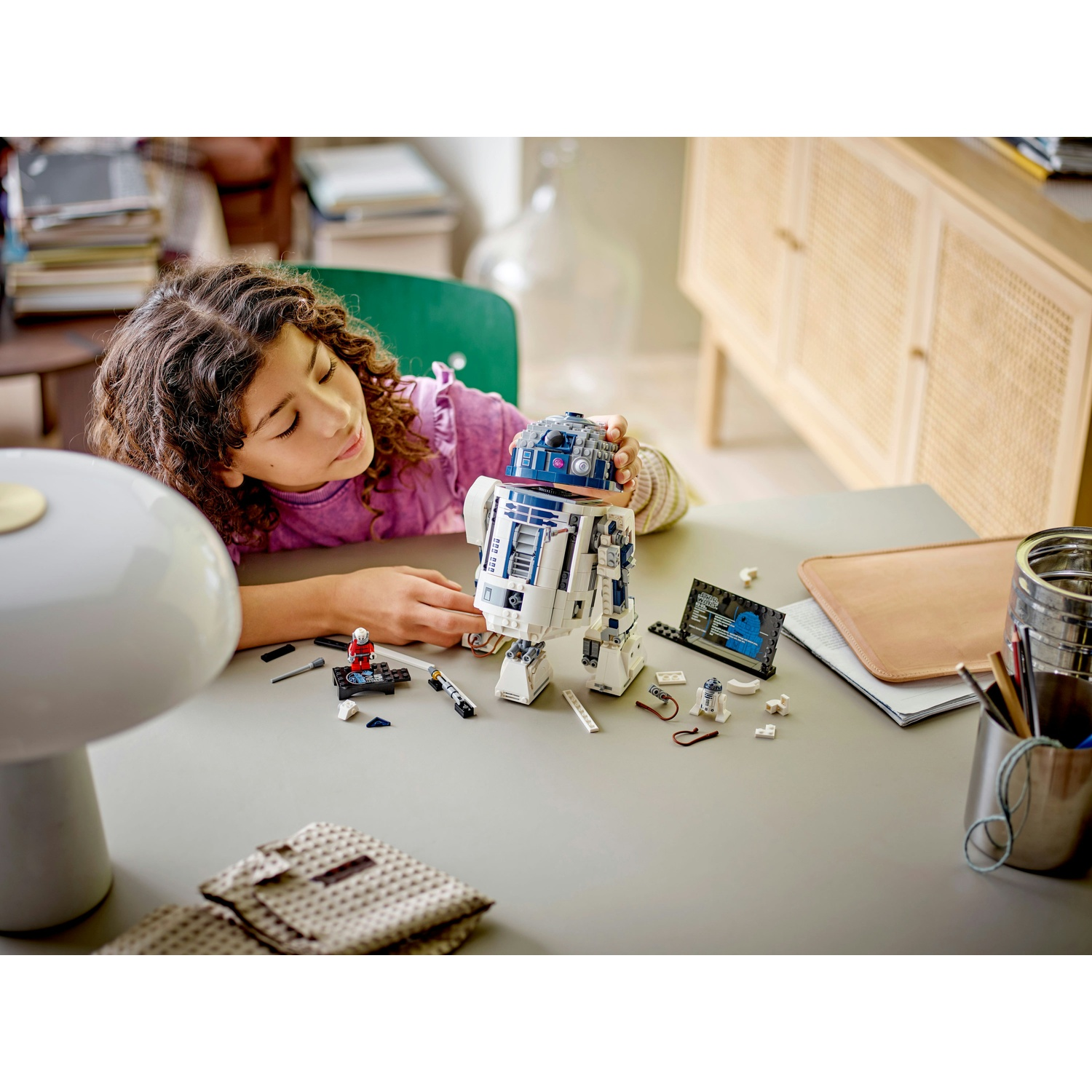Una ragazza esamina un set Lego R2-D2 di Star Wars su un tavolo con altri pezzi Lego, una penna e un blocco note.