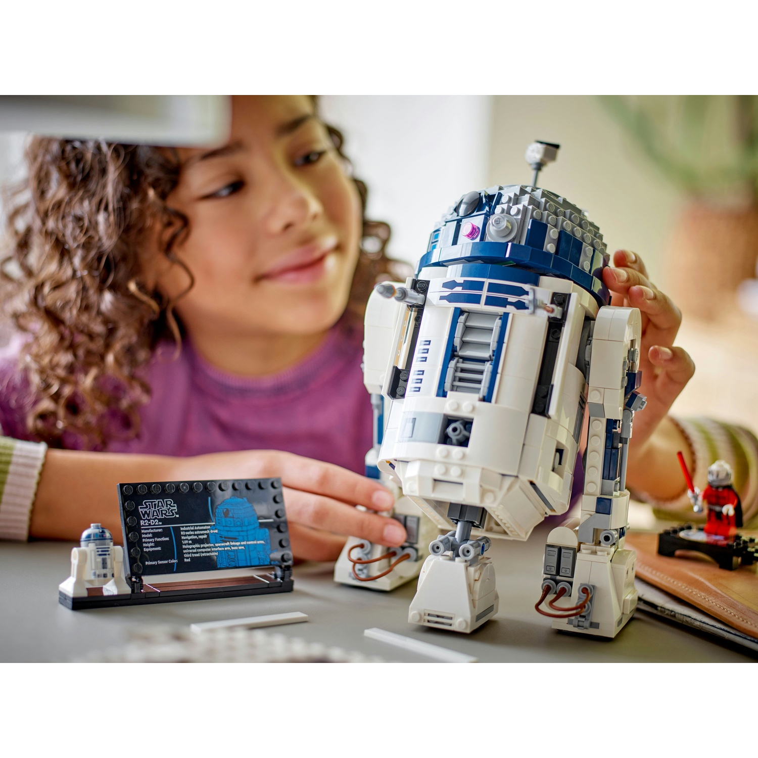 Una ragazza che costruisce Lego R2-D2, con il modello e una scheda espositiva, una figura rossa vicina.