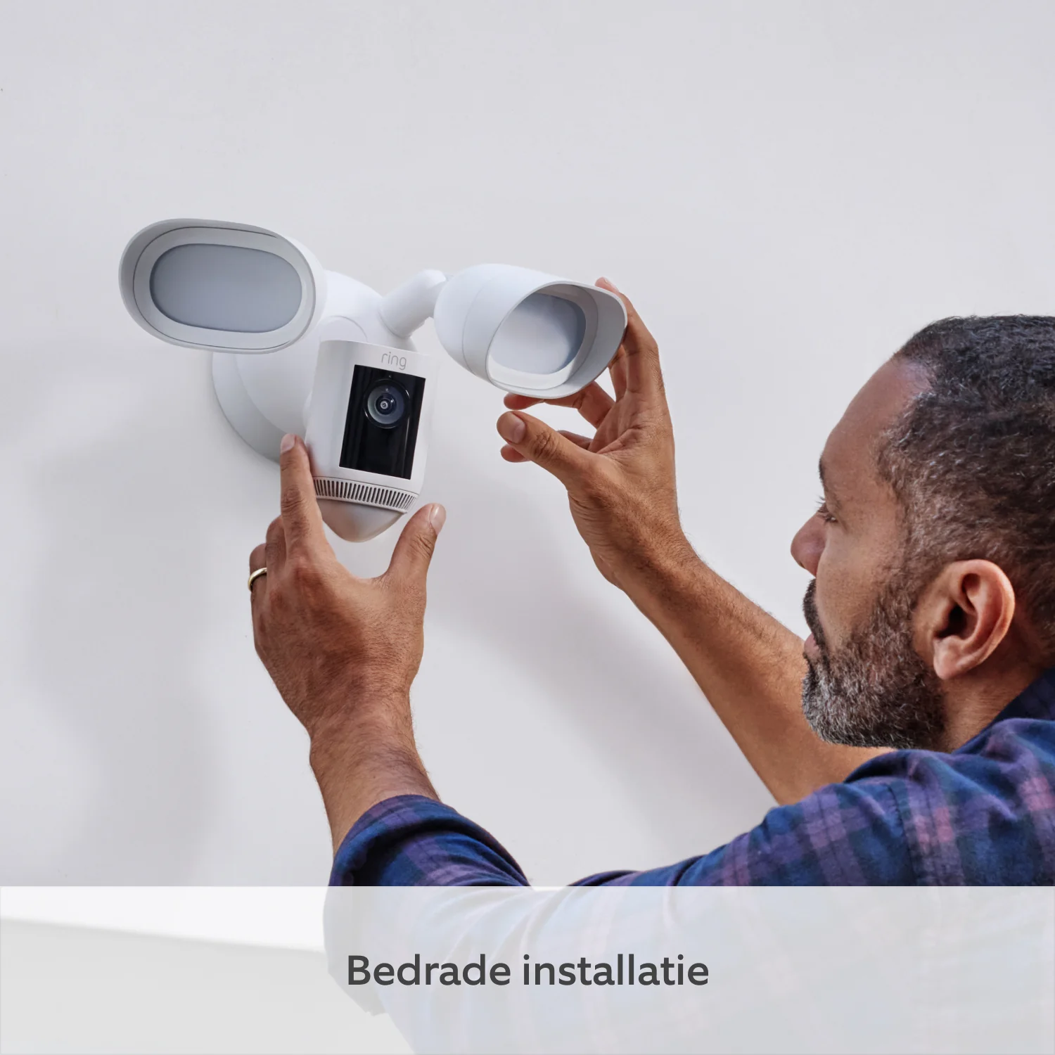 A man installs a white security camera on a white wall. He is wearing a blue plaid shirt.