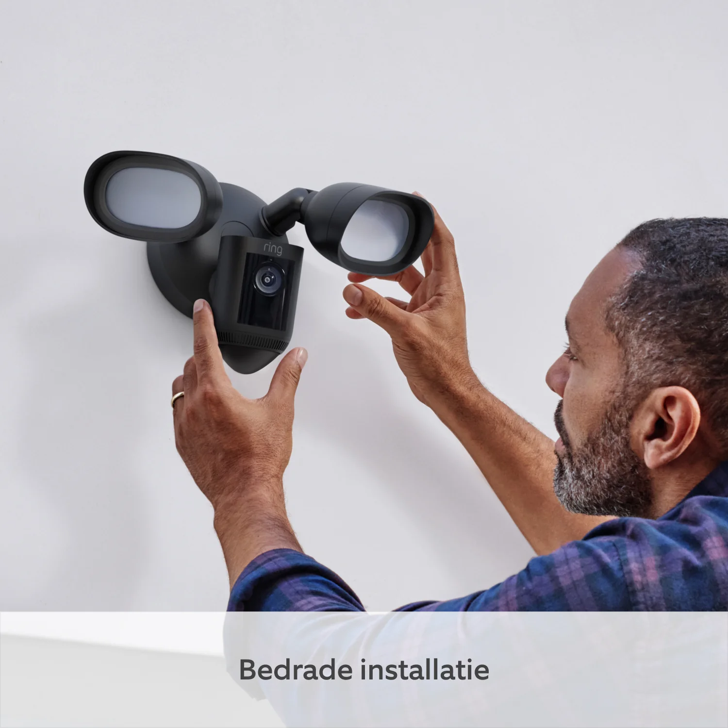Man installeert een zwarte Ring schijnwerpercamera op een witte muur. Tekst: Bedrade installatie.