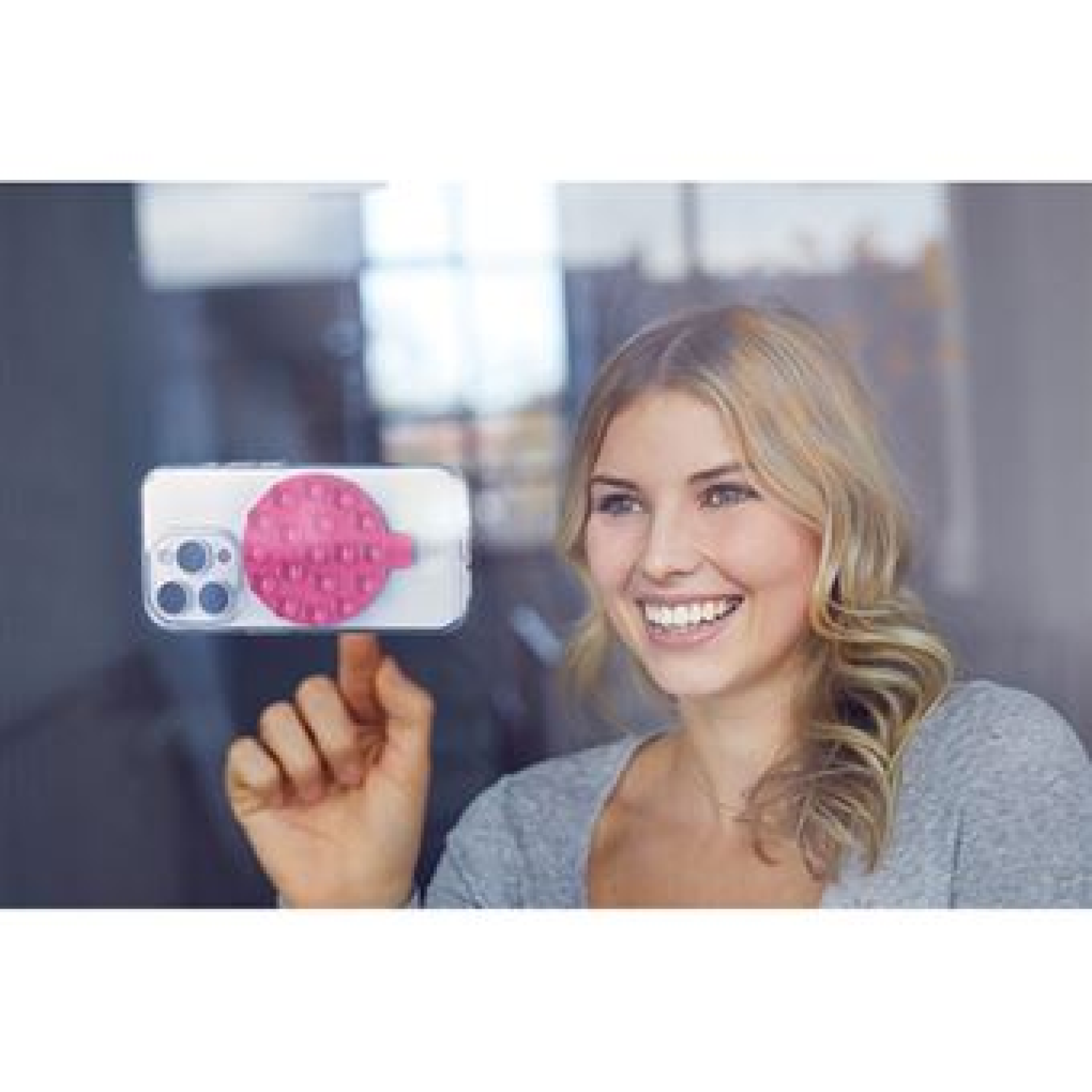 Una mujer sonríe, sosteniendo un teléfono con un pop socket rosa contra una ventana.