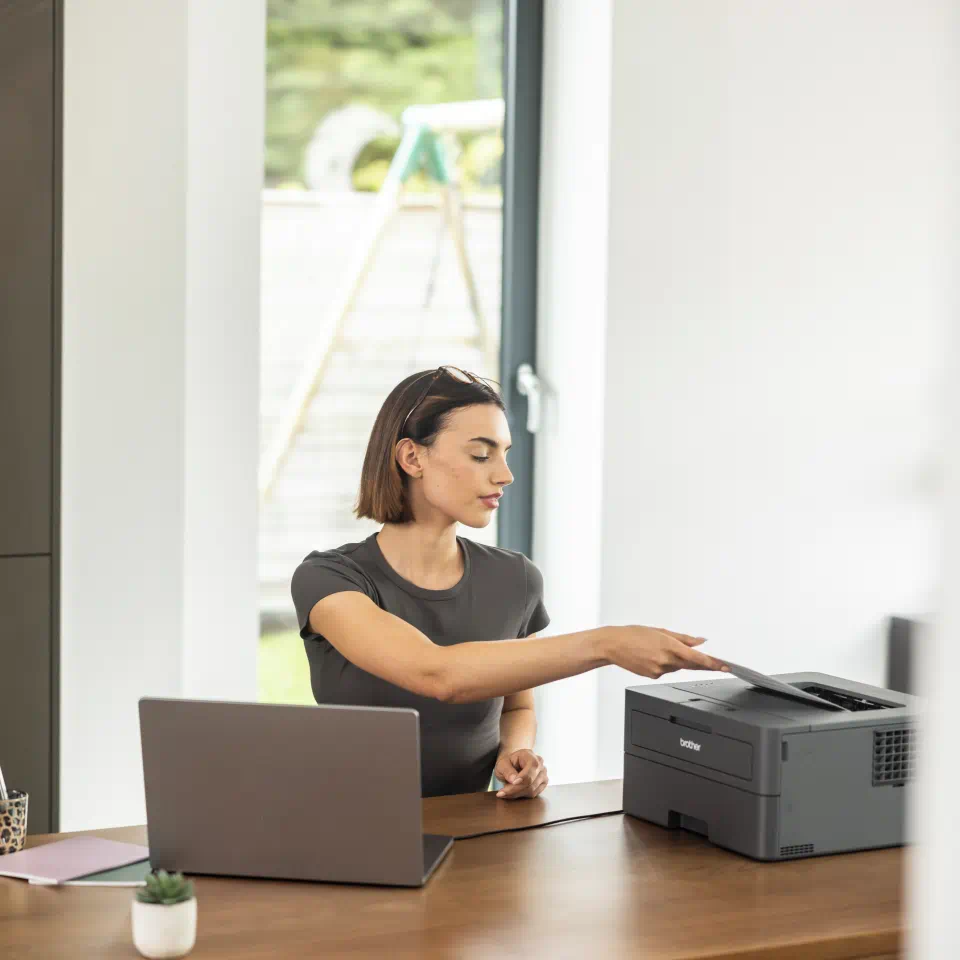 Nő az asztalnál, dokumentumot nyomtat laptopról. A nyomtató az asztalon van.
