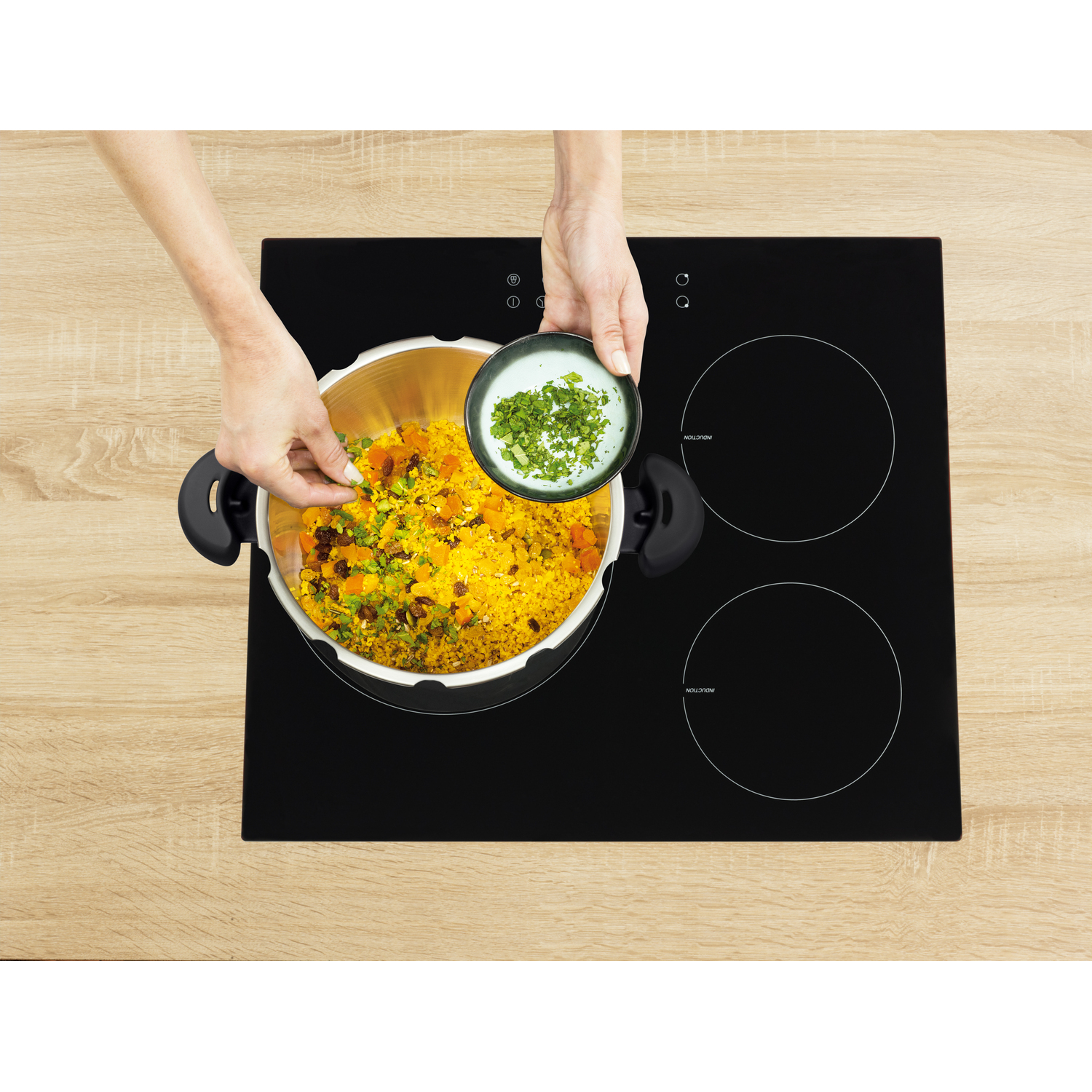 Hands adding garnish to food in a pot on a black induction cooktop.