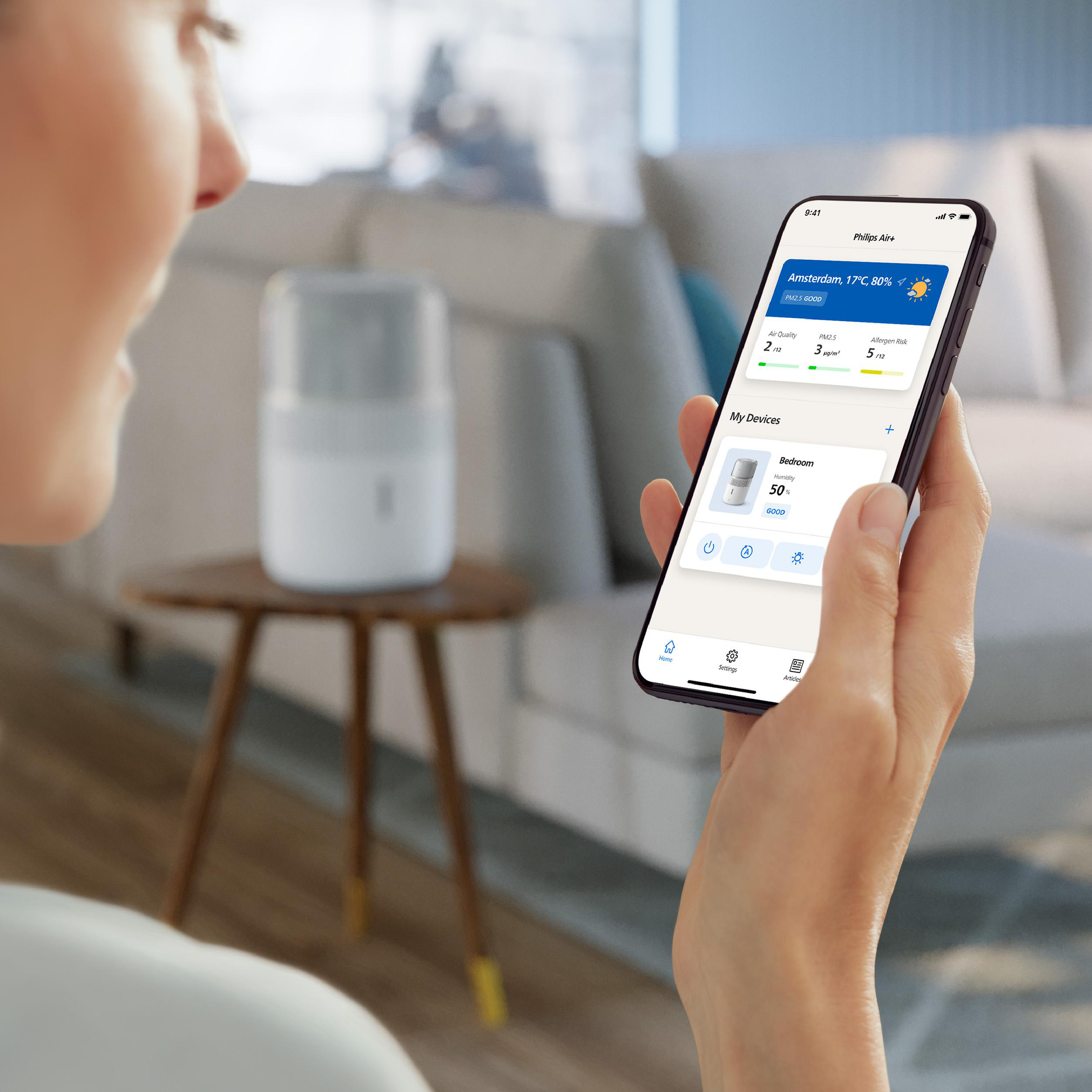 Frau hält Telefon mit App. Luftreiniger auf Tisch im Hintergrund. Sofa und heller Raum.