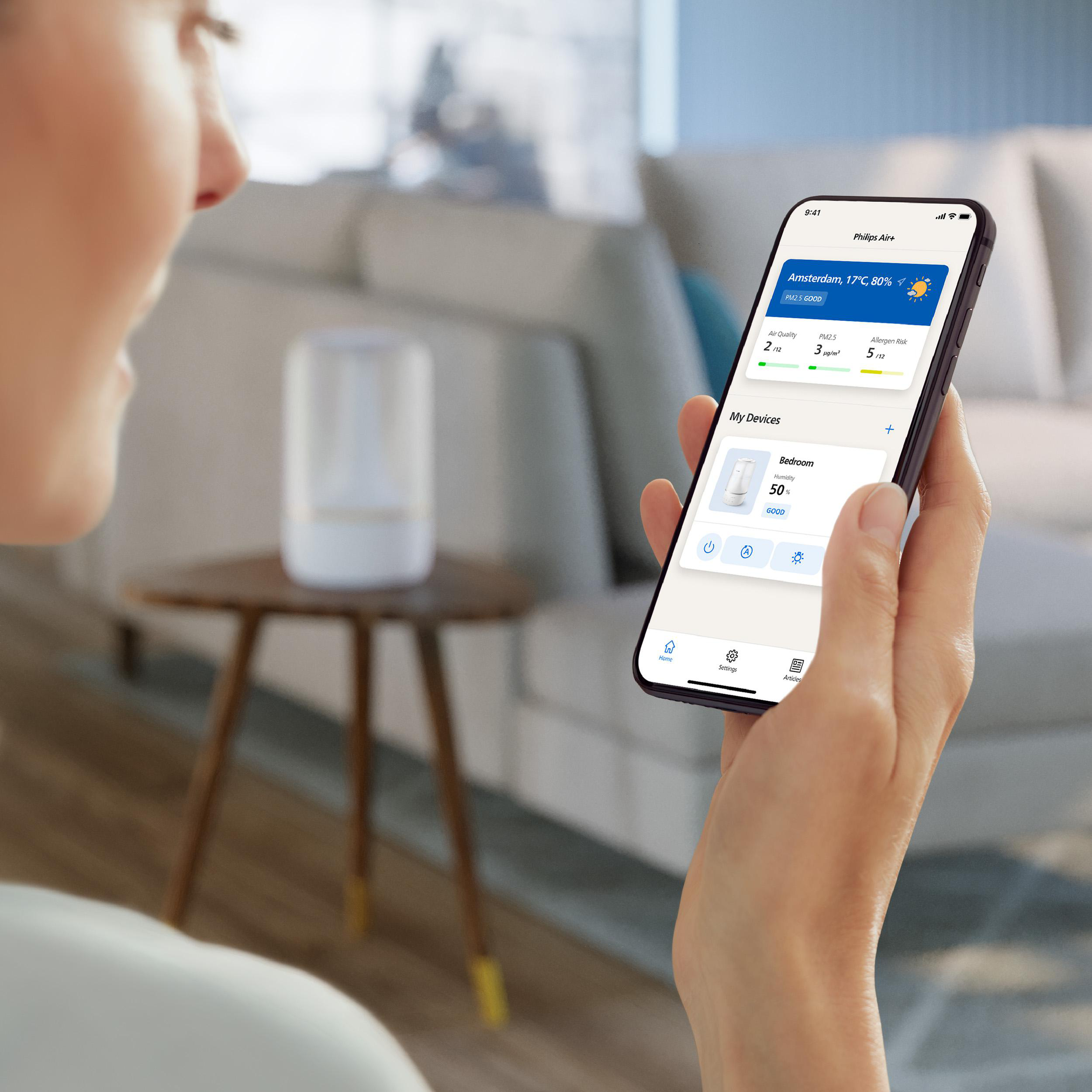 Eine Person hält ein Smartphone, das eine Smart-Home-App zeigt. Ein weißer Luftbefeuchter ist im Hintergrund.