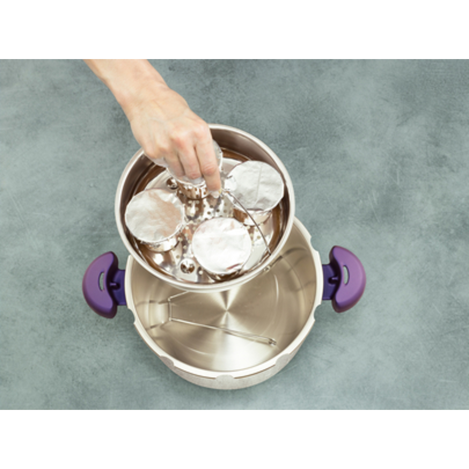 A hand places a tray with covered food containers into a stainless steel steamer pot.