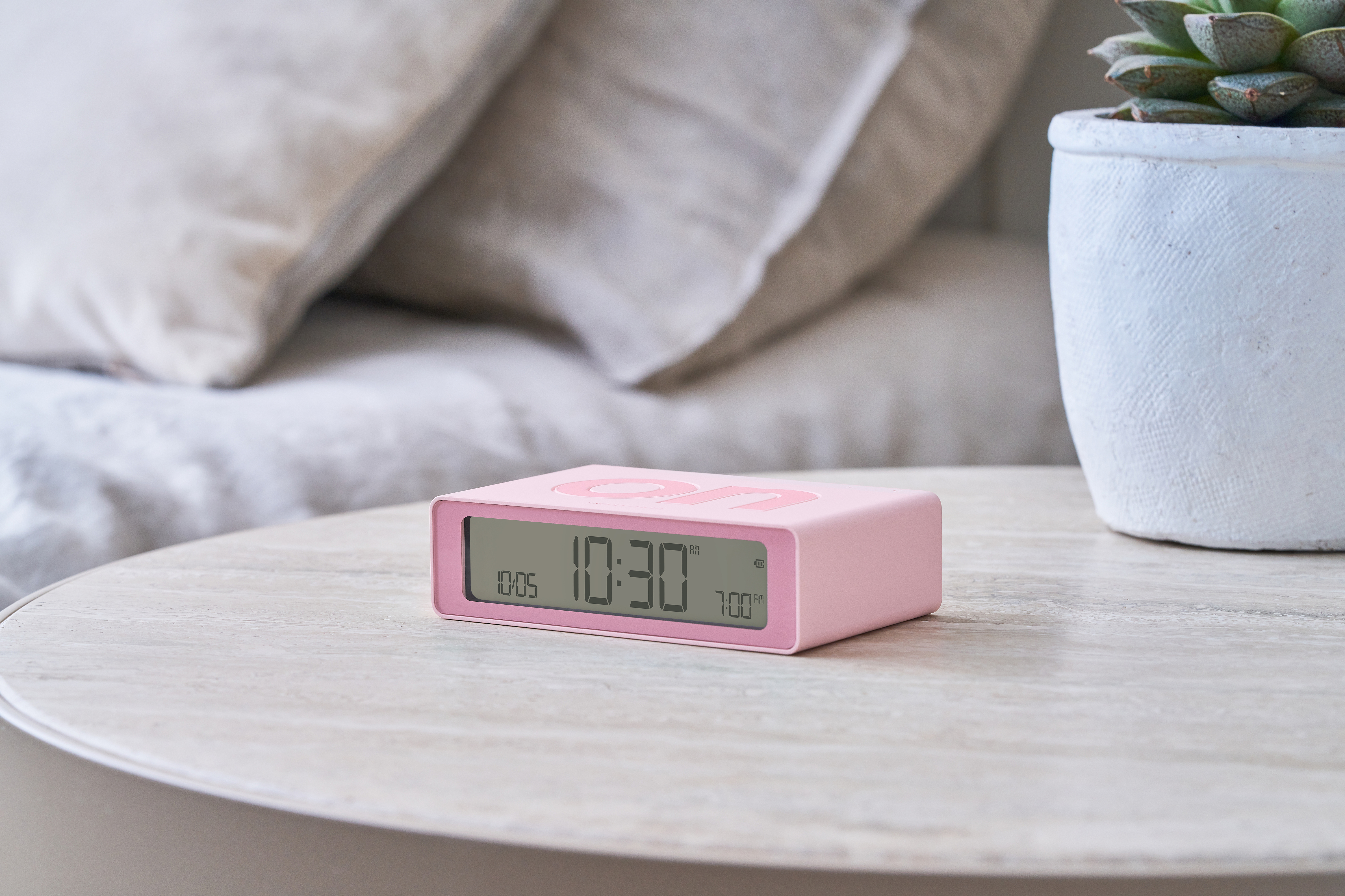 Un orologio a ribalta rosa mostra le 10:30. È su un tavolo con una pianta e cuscini.