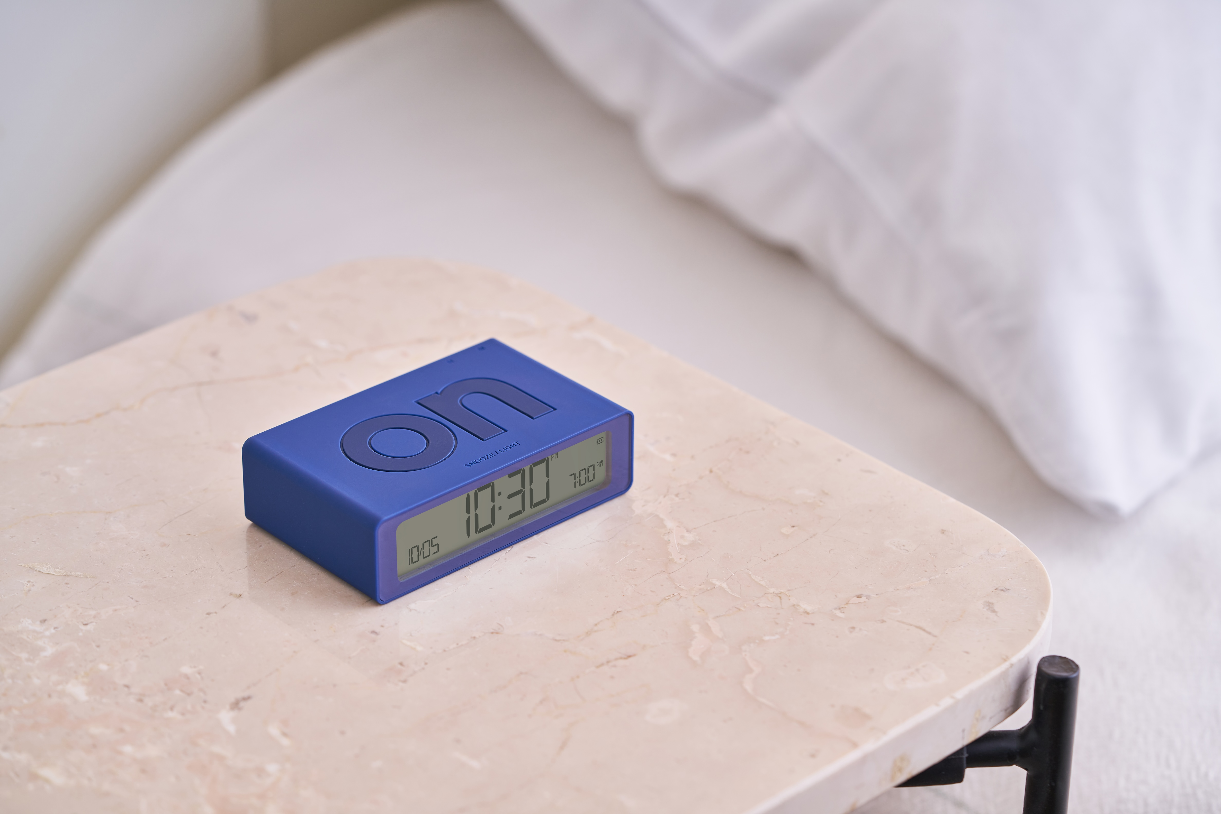 Sveglia blu su un comodino di marmo accanto a un letto bianco con un cuscino.