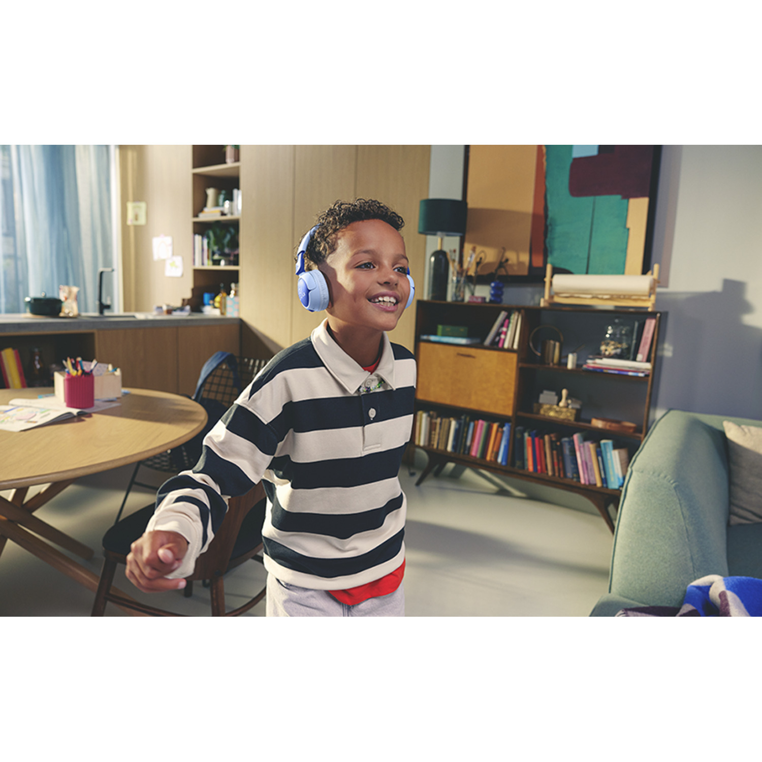 Garçon avec casque bleu sourit. Arrière-plan: salon avec bibliothèque et table.