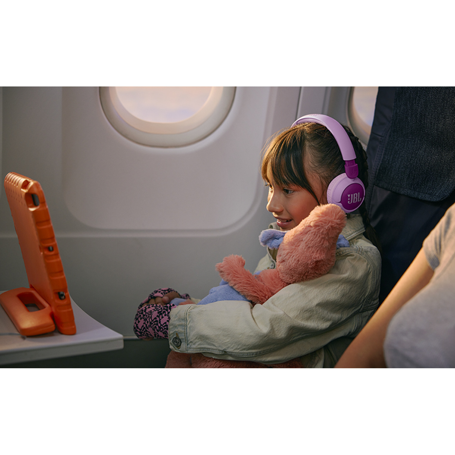 A child with headphones watches a tablet in an airplane with a pink plush.