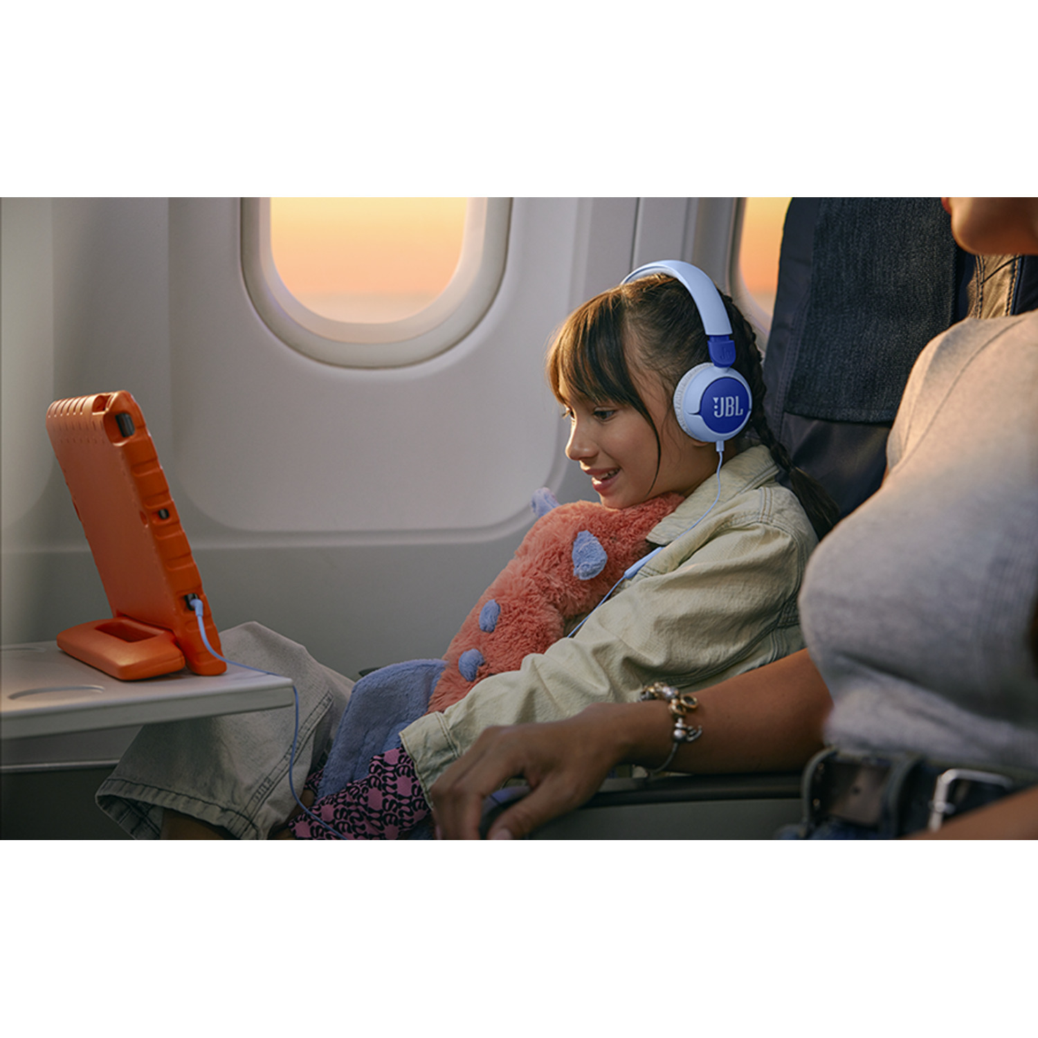 A girl with headphones watches a tablet on a plane. An adult's arm is in the foreground.