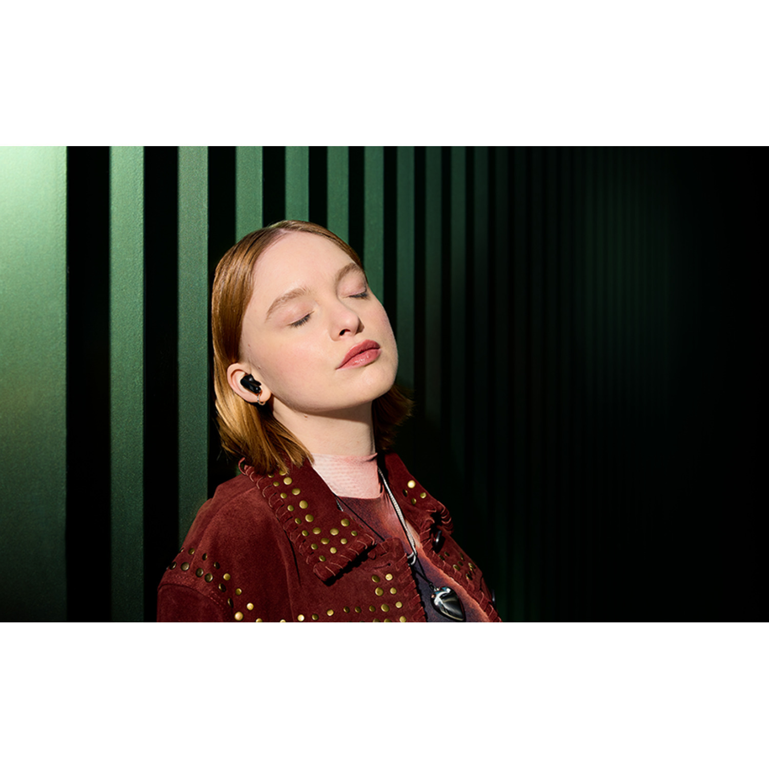 Woman with closed eyes wearing earbuds, red jacket.  Green vertical striped background.