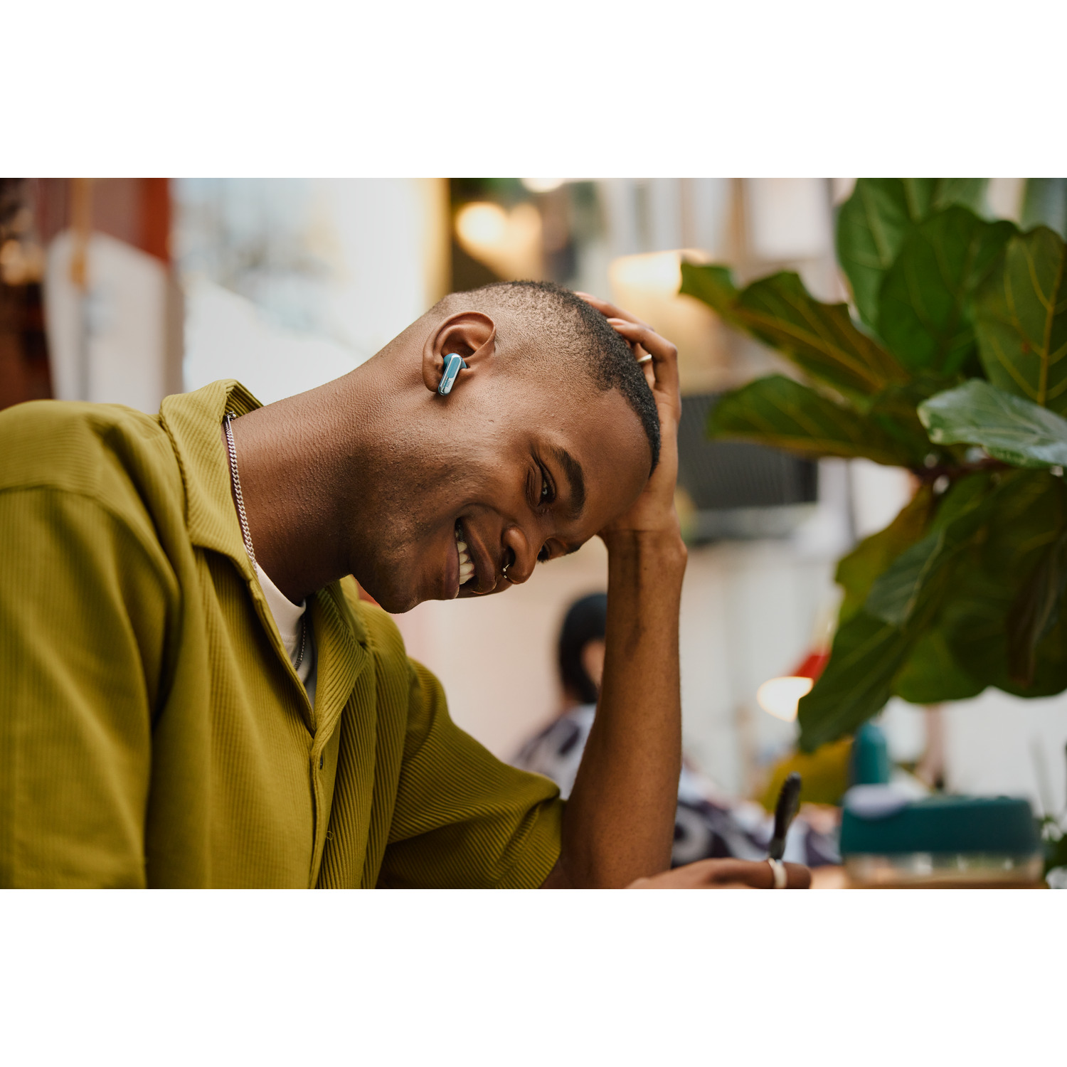 Smiling man with earbuds. He wears a yellow shirt and has a nose piercing. Blurred background.