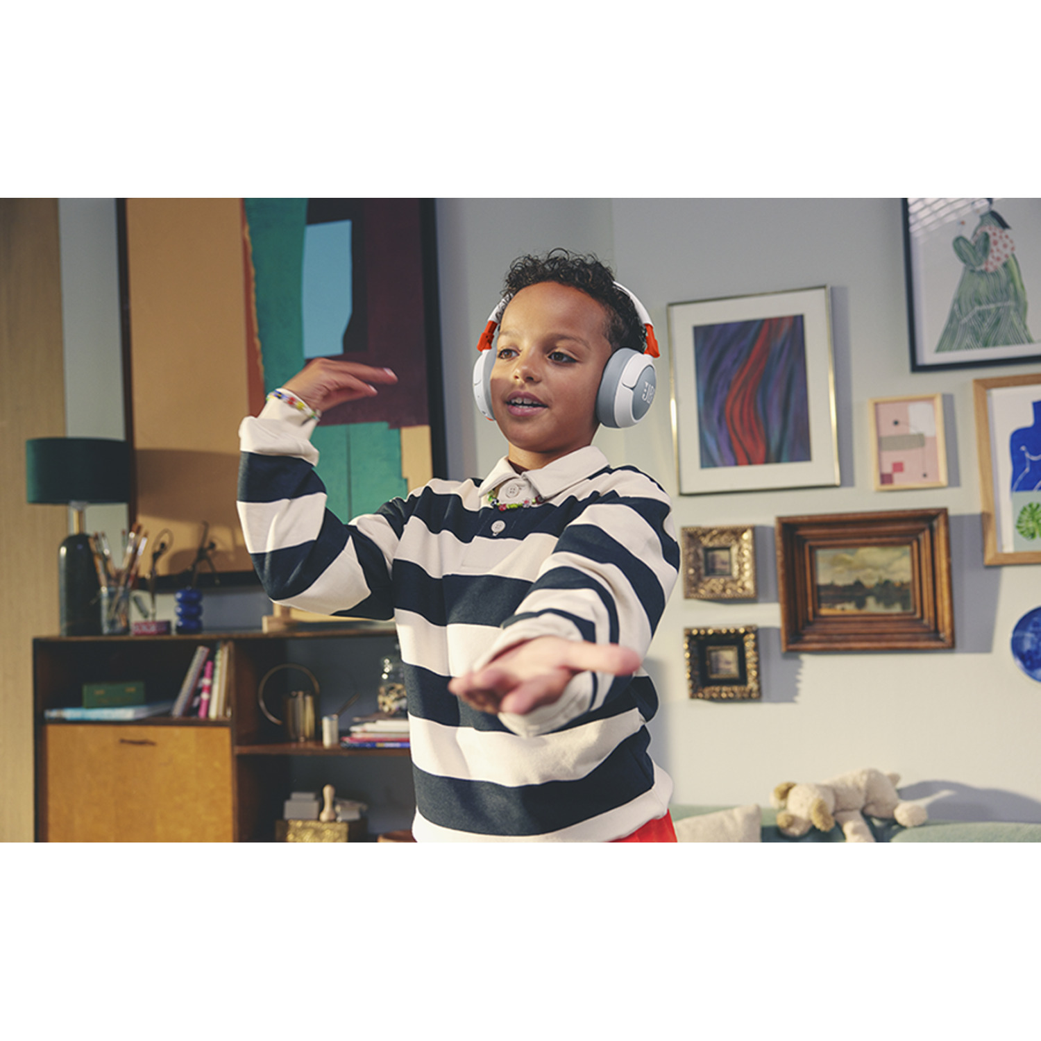 A young boy wearing striped shirt and headphones, gesturing with hands, surrounded by art and furniture.
