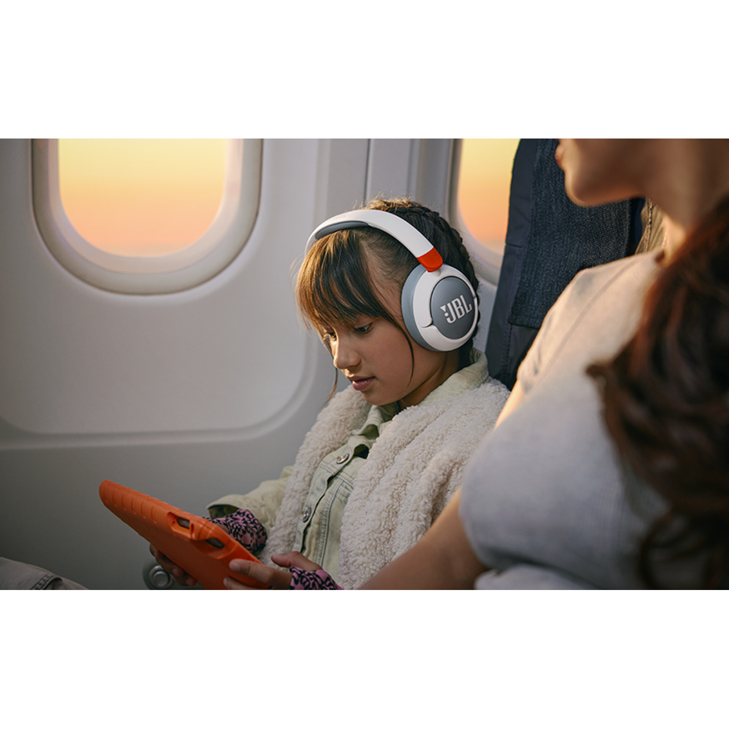 Girl wearing headphones on a plane. Tablet in hand. Sunset visible through window.