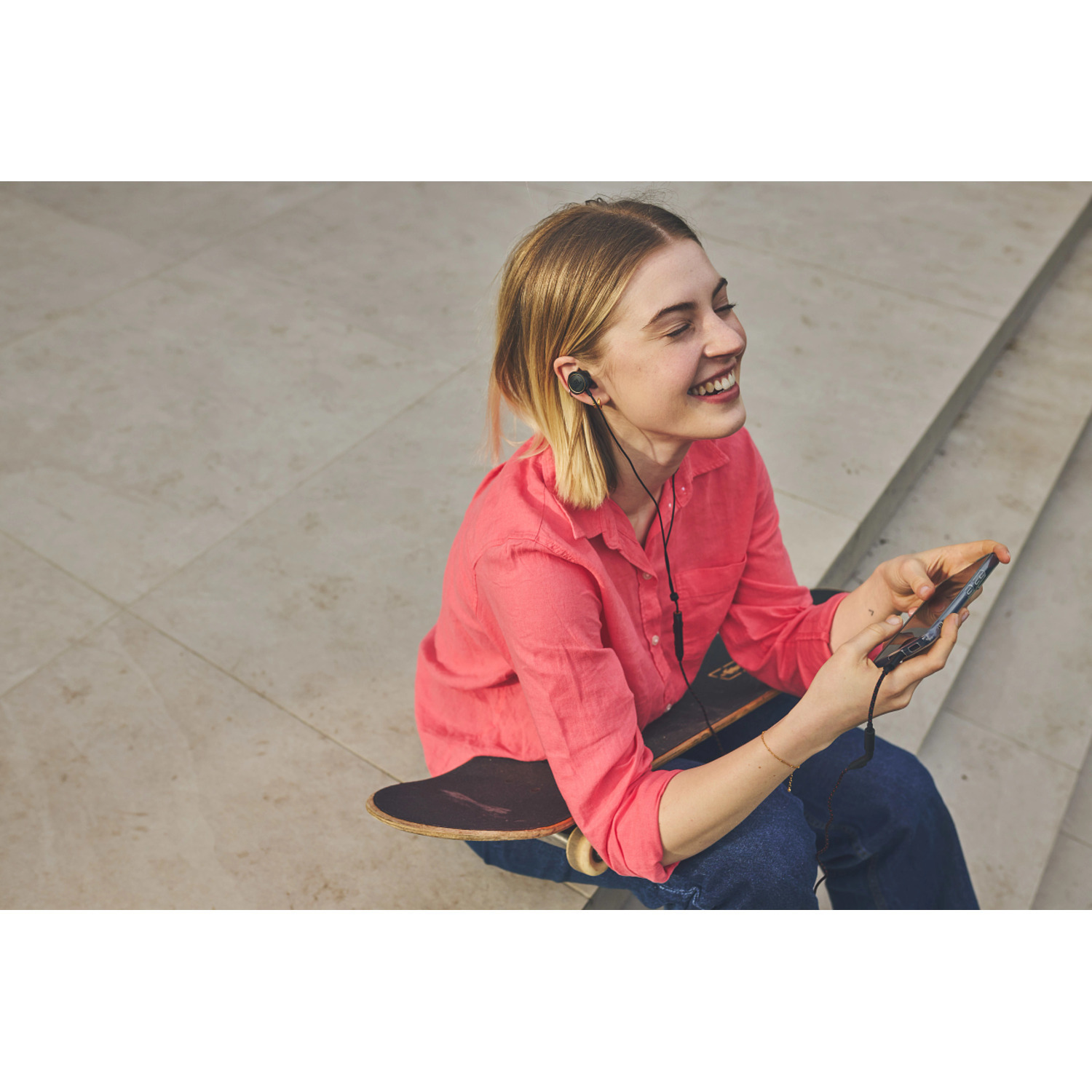 Femme aux cheveux blonds, chemise rose, avec des écouteurs, souriante, tenant un téléphone près d'un skateboard.