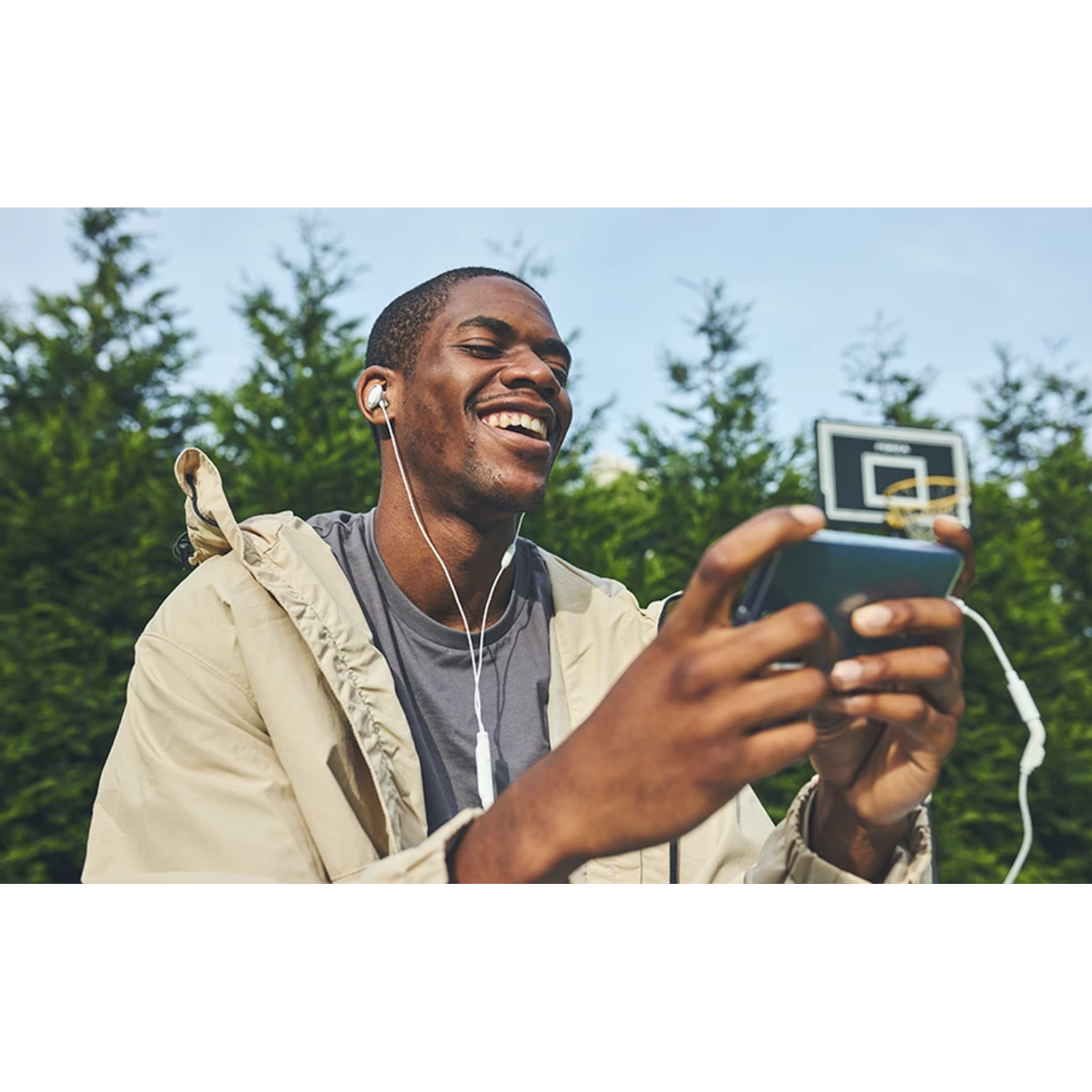 Un homme souriant tenant un téléphone, avec des écouteurs, devant un terrain de basket.