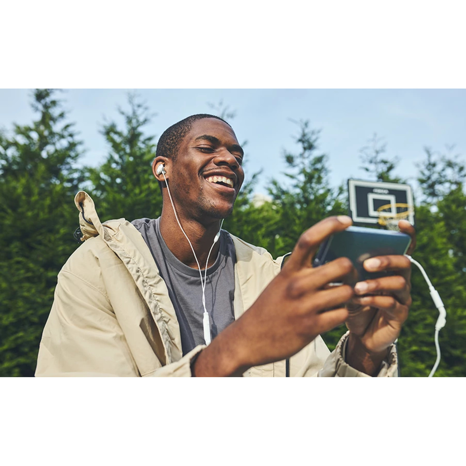 Un homme avec des écouteurs sourit en utilisant un smartphone. Un panier de basket est en arrière-plan.