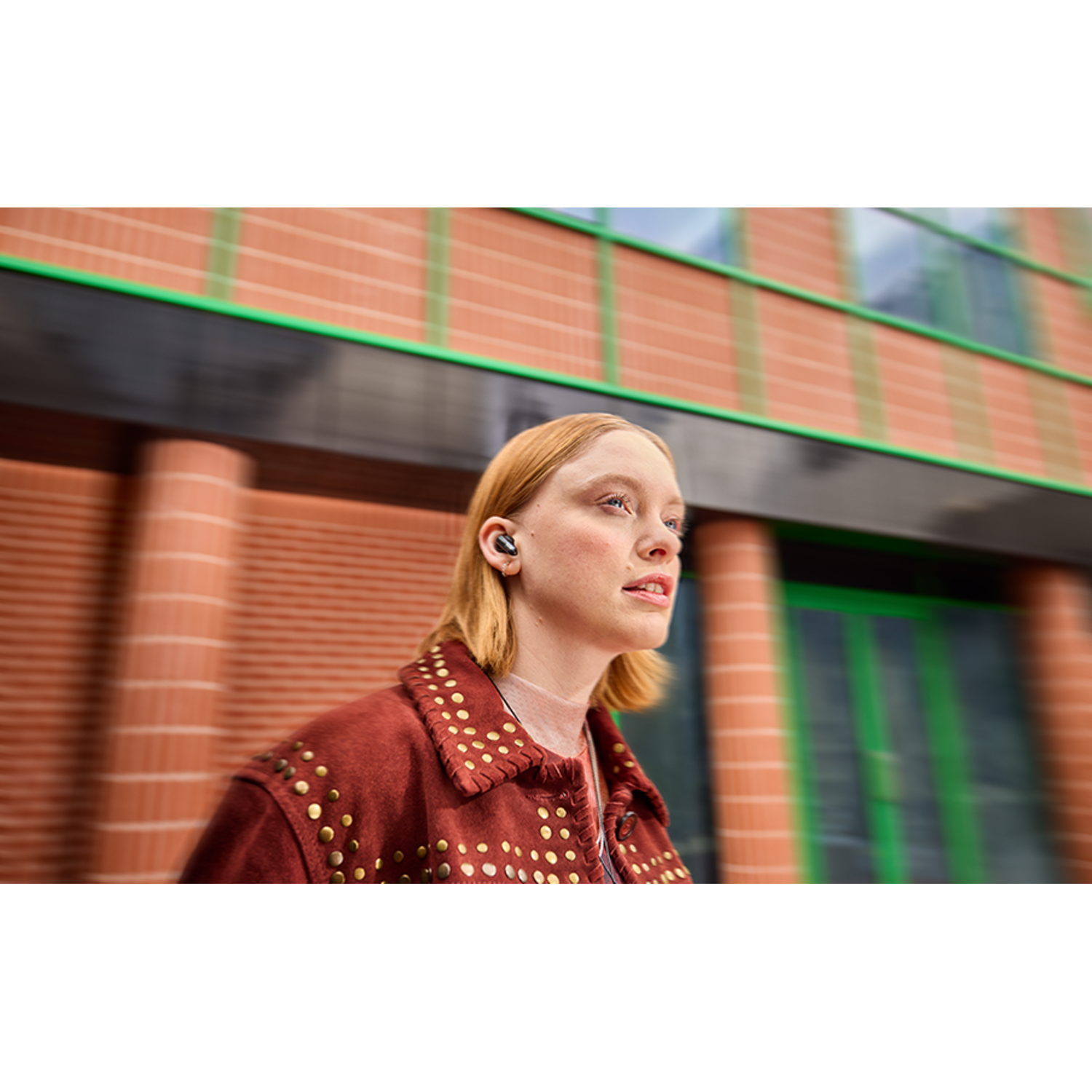A woman with blonde hair wearing earbuds. She wears a red jacket with gold studs.