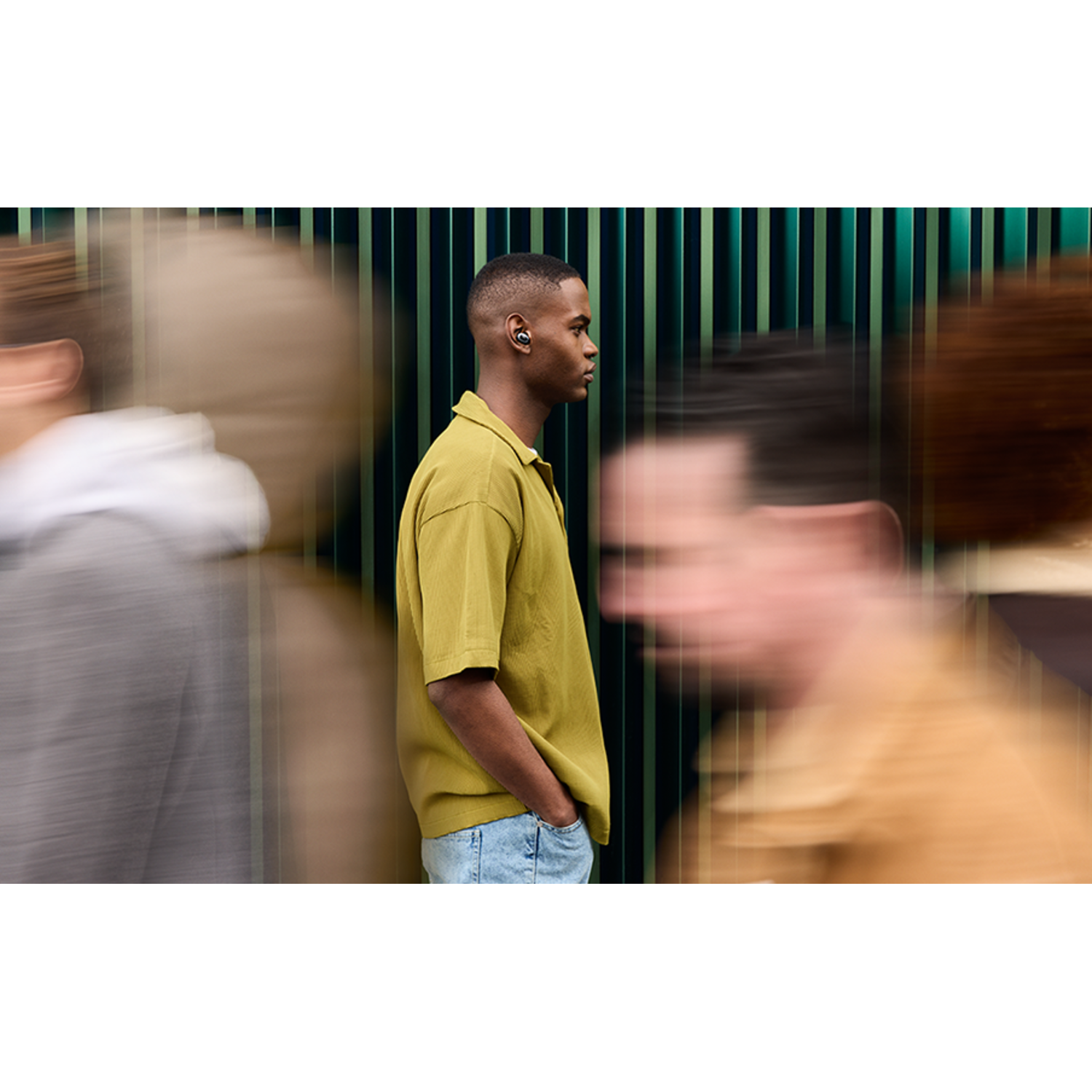 A man wearing earbuds stands with hands in his pockets, in front of blurry figures.