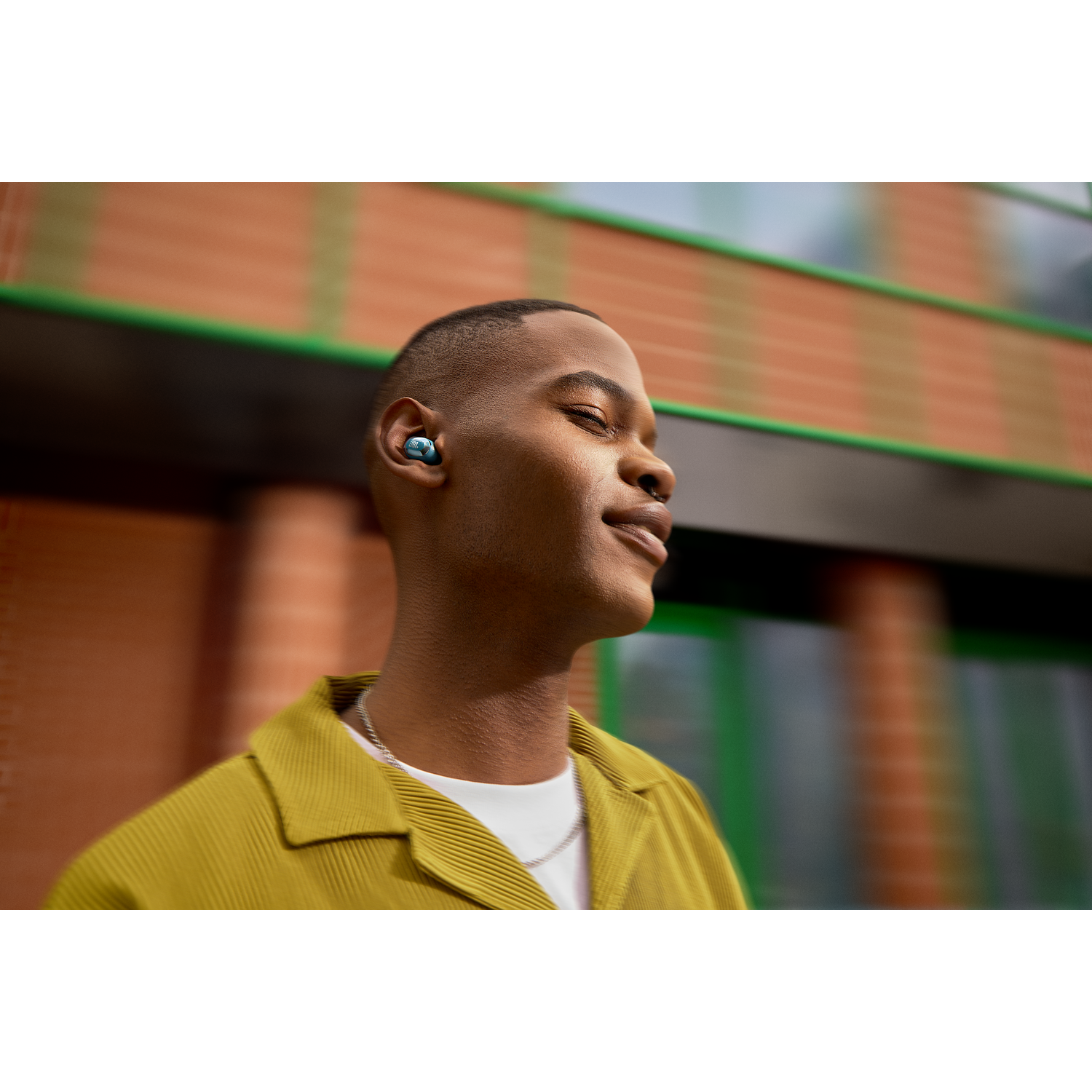 A man wearing earbuds smiles outdoors. Earbuds are blue. He wears a yellow shirt and has a chain.