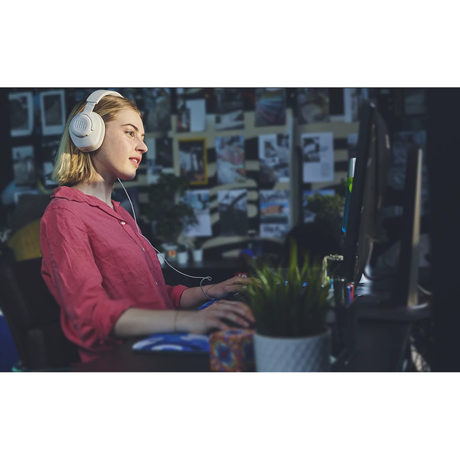 Woman wearing white headphones, working on a computer. Desk with a plant and a monitor.