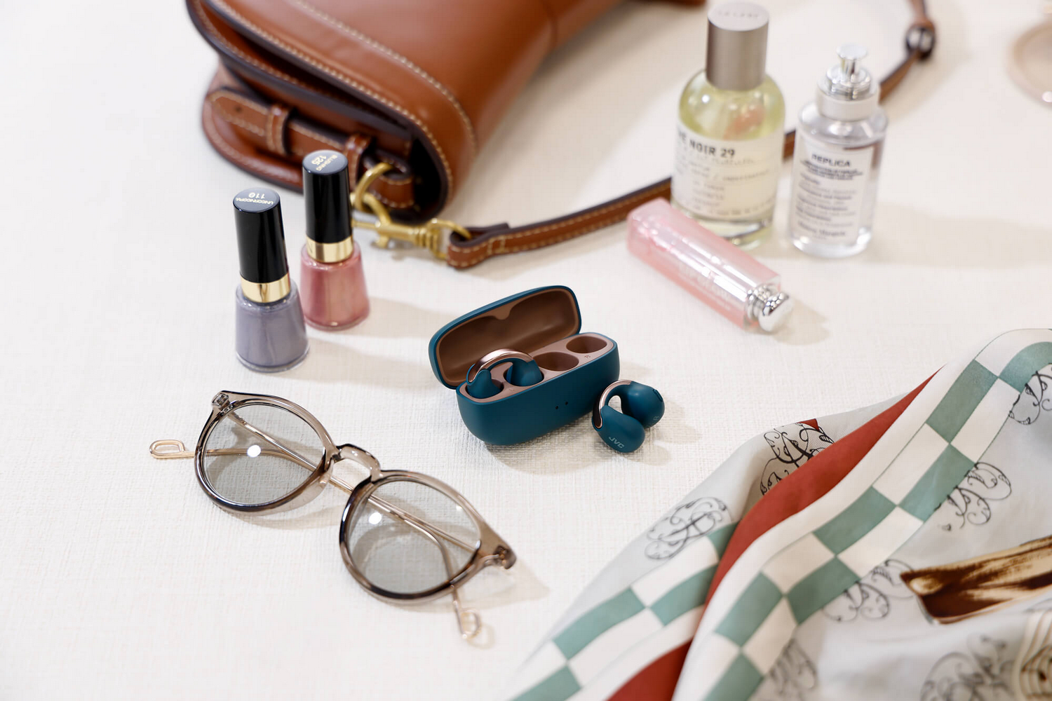 Varios objetos sobre una mesa blanca: bolso, perfumes, esmalte de uñas, gafas de sol y auriculares.