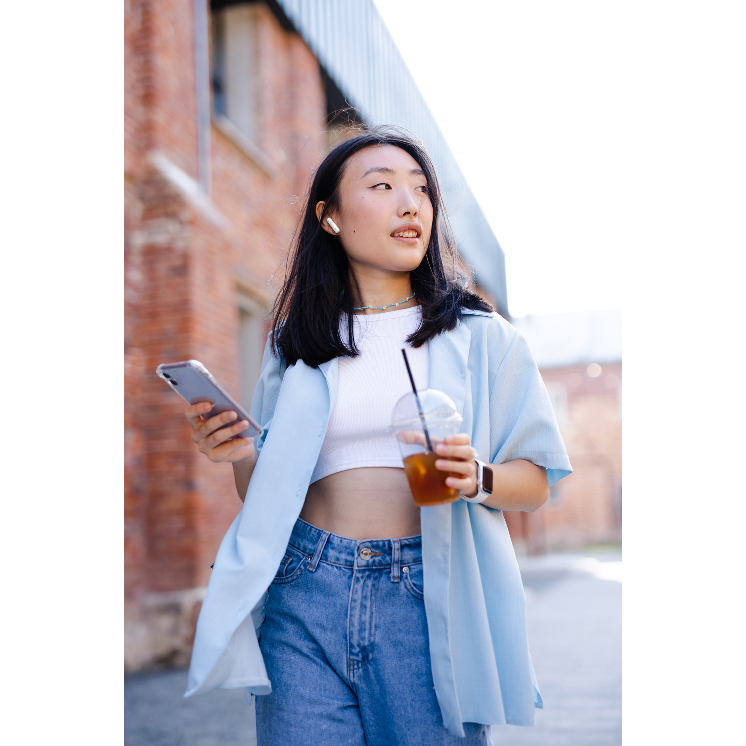 A woman holding a phone and a drink, walking by a brick building, wearing earbuds.