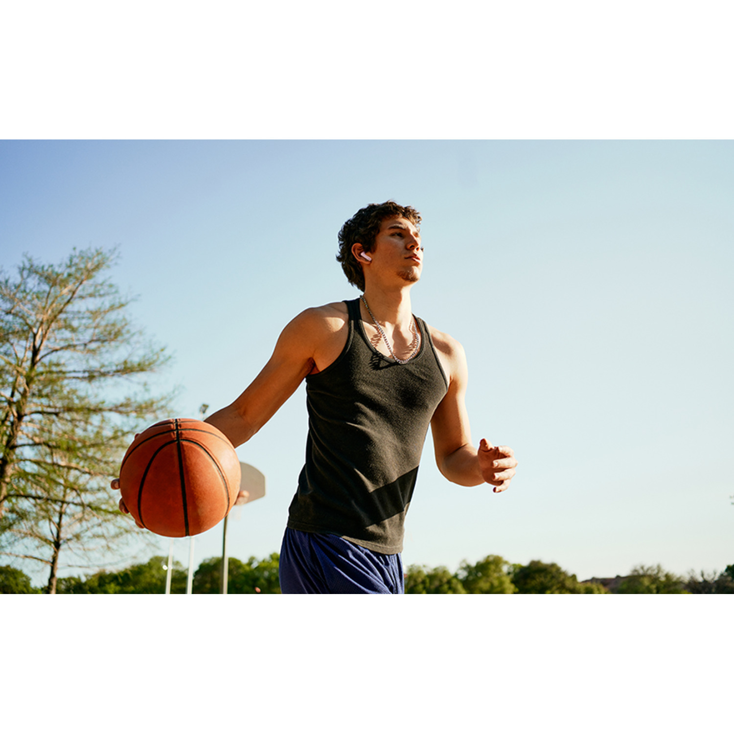 A man in a black tank top dribbling a basketball on a court, with trees and a clear sky in the background.