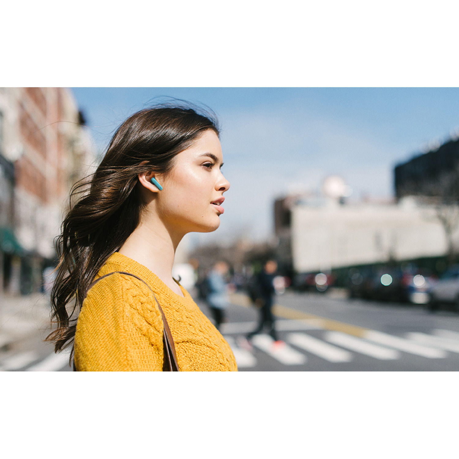 Woman with yellow sweater, wearing teal earbuds outdoors in a city.