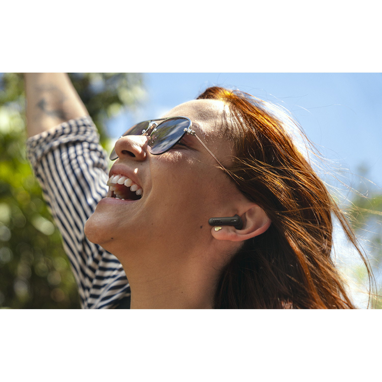 A woman with sunglasses and an earbud smiles, looking up at the sky.