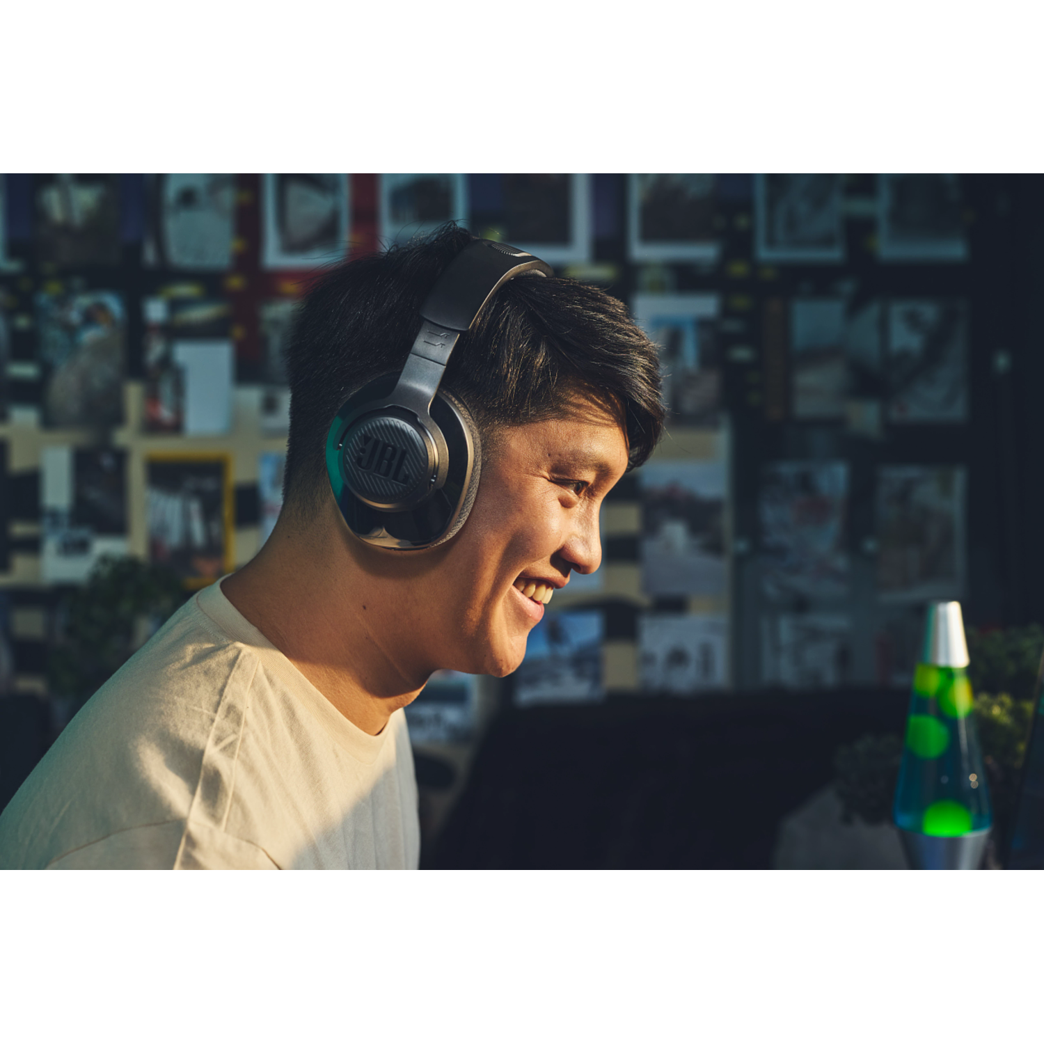 Homme portant un casque JBL, souriant. Arrière-plan flou, avec une lampe à lave et d'autres objets.