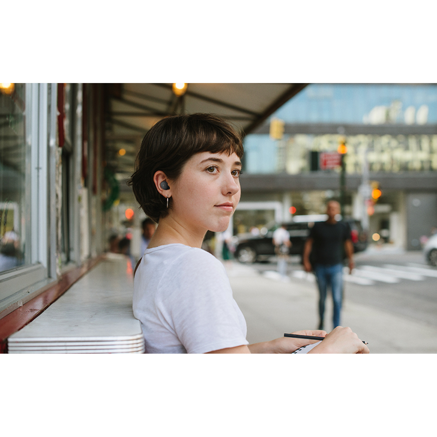 A young woman with earbuds in, looking away, sitting at a table on a city street.