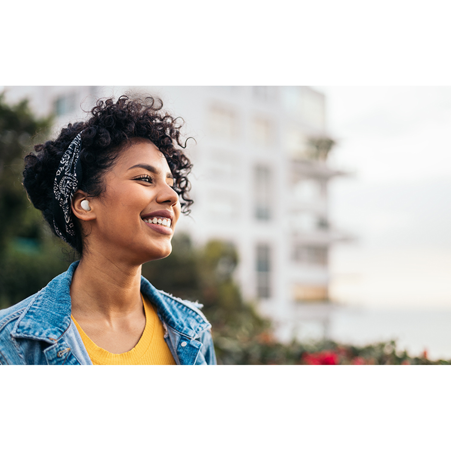 Woman with earbuds smiling, wearing a denim jacket. Background is a city view.