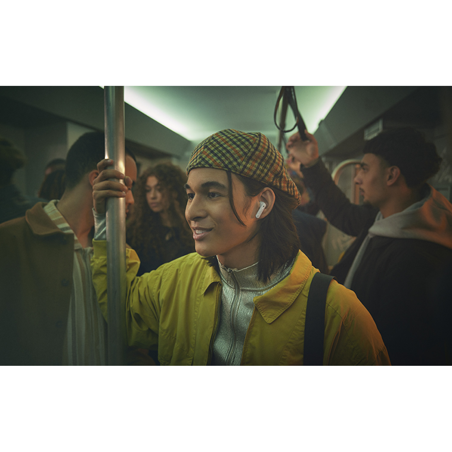 Man on a train wearing earbuds. People in the background. Wearing a yellow jacket and a cap.