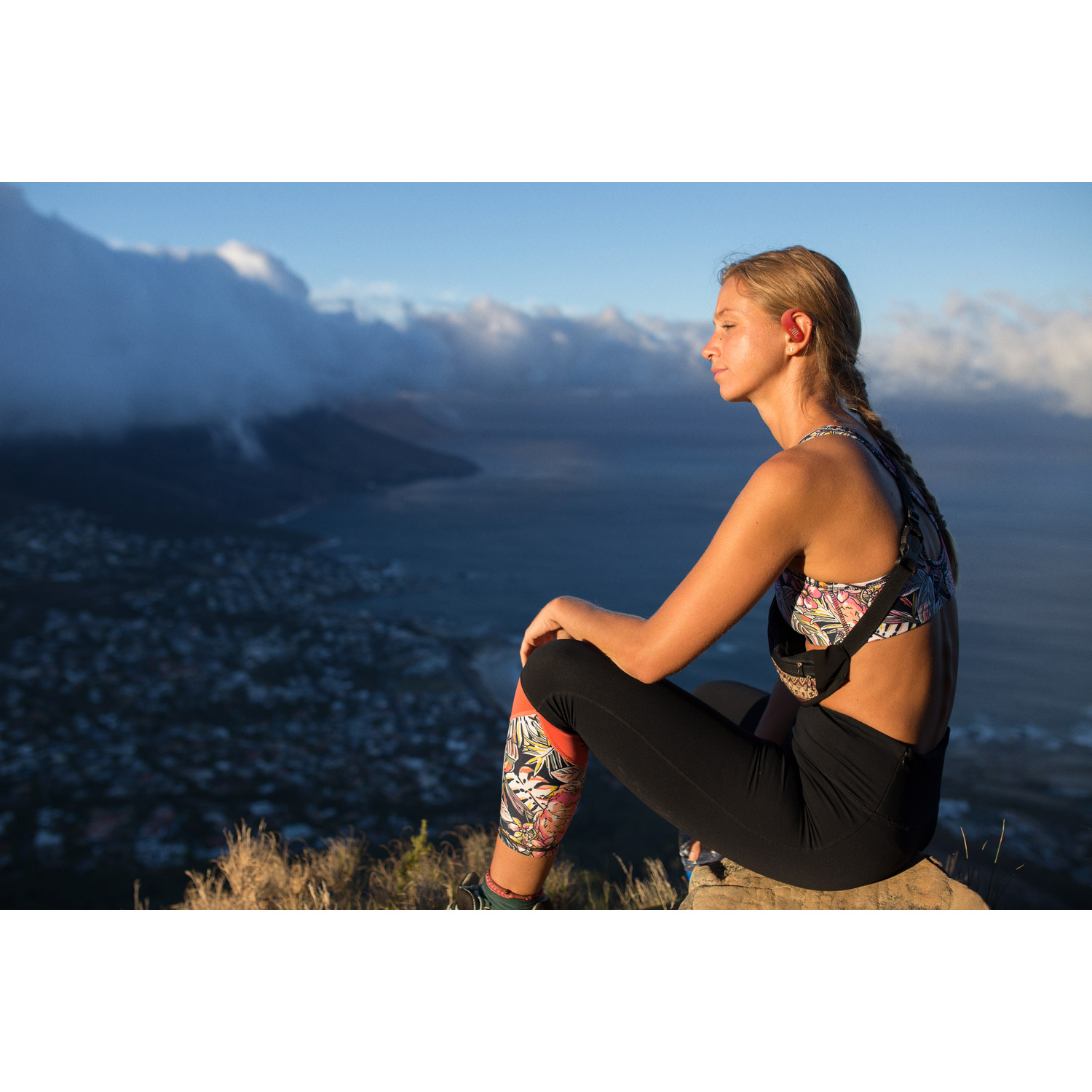 Woman sitting on a mountain with a view. She wears a sports bra and leggings.