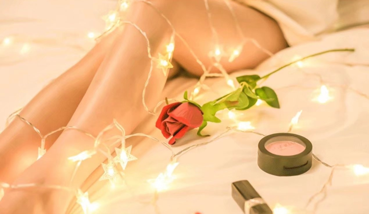 Nogi osoby na łóżku z lampkami choinkowymi, różą, pudrem do makijażu i szminką.