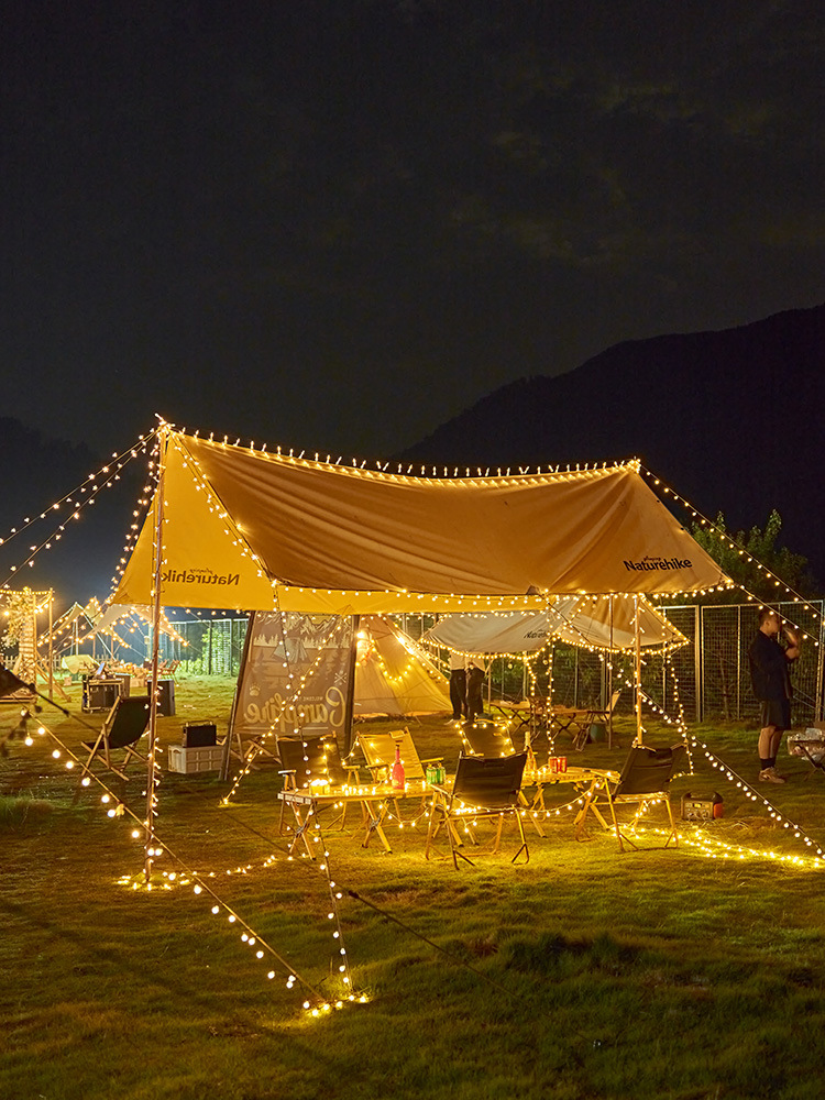 Kemping z rozwieszonymi światłami nad namiotami i stołami w nocy, z górami w oddali.