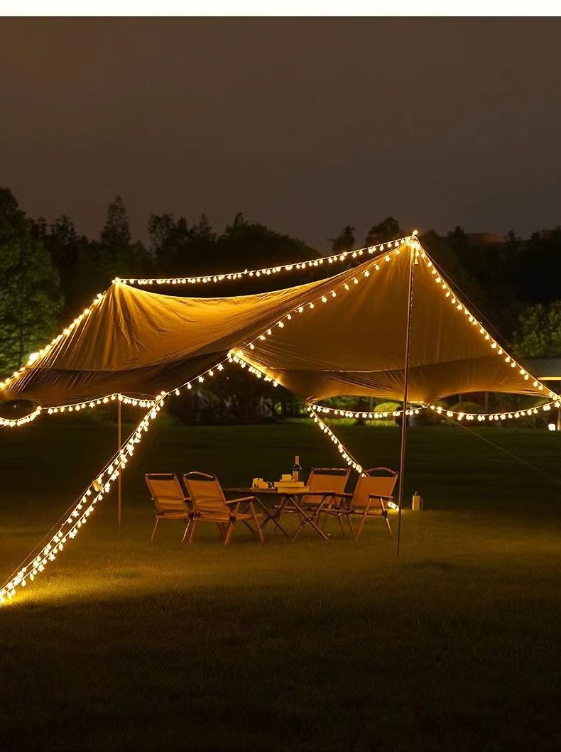 Scena na zewnątrz z namiotem oświetlonym lampkami, stołem, krzesłami, w nocy.